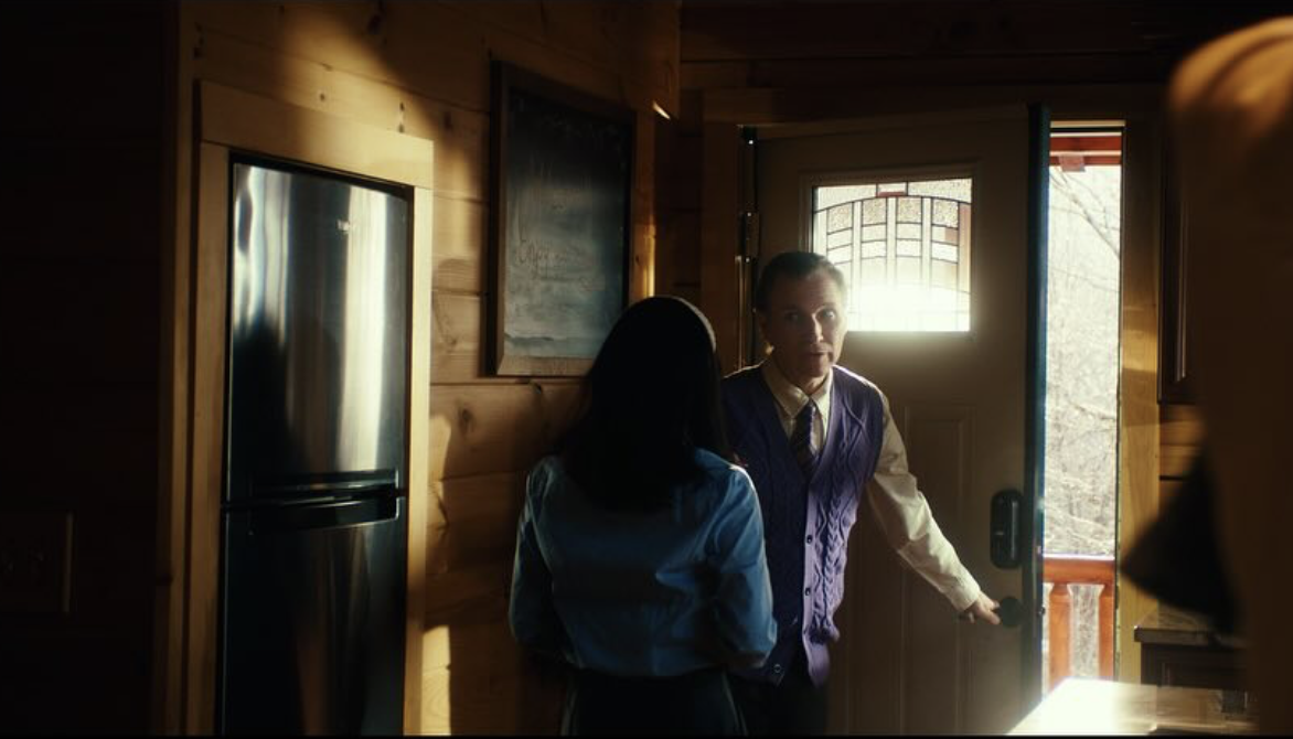 A woman with black hair and a blue jacket faces a man in a purple vest and white shirt at the door of a wooden cabin, with sunlight coming through the window.