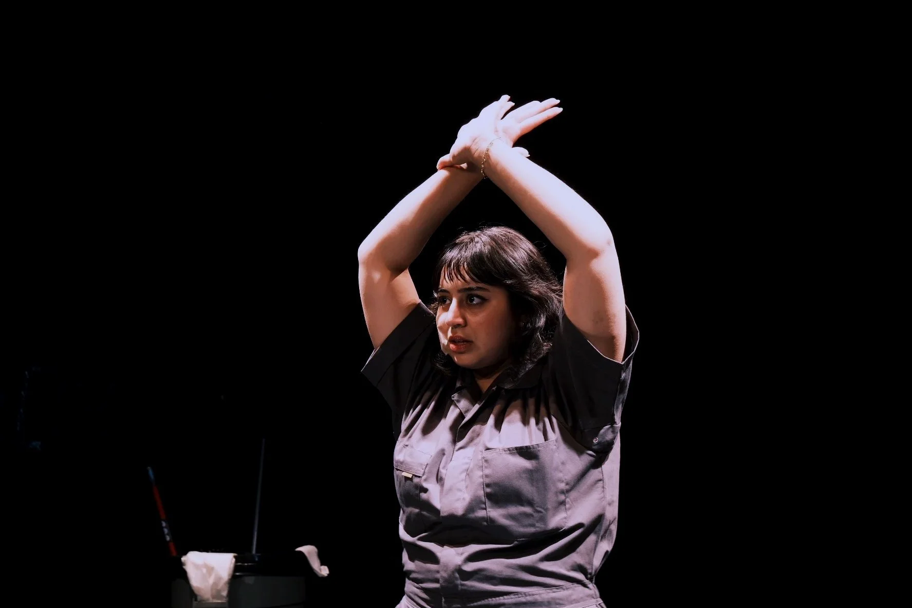 A woman with dark hair and wearing a uniform appears to be in a performance or rehearsal, with her arms raised above her head crossing at the wrists against a black background.