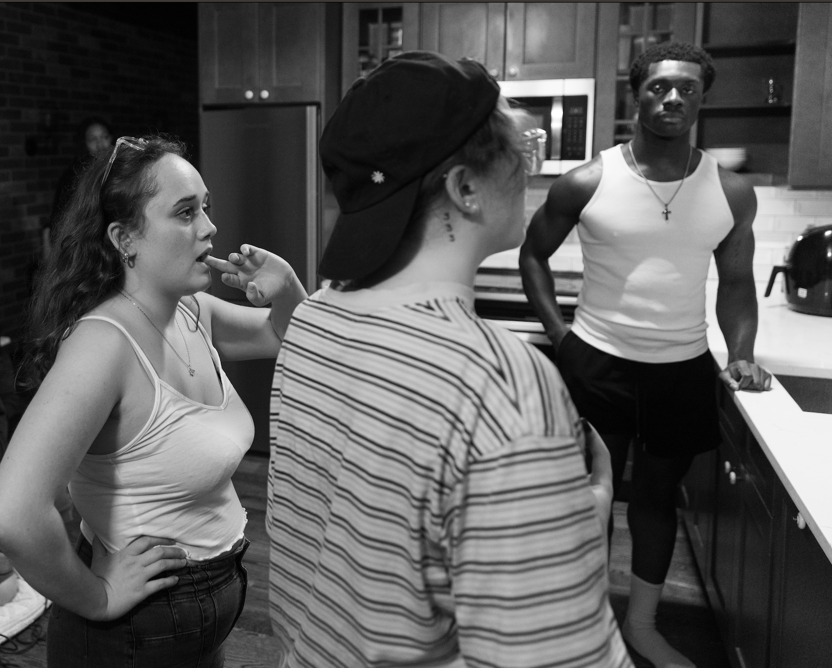 Four young adults, two women and two men, are gathered in a kitchen having a conversation. The woman on the left is wearing a sleeveless top and has her hand on her hip, listening attentively. The woman in the middle is wearing a striped shirt and a backwards cap, facing the other three. The man on the right, who is muscular, is wearing a white tank top and a cross necklace, standing with his arms on the kitchen counter, paying attention to the group. The kitchen has wooden cabinets and stainless steel appliances.