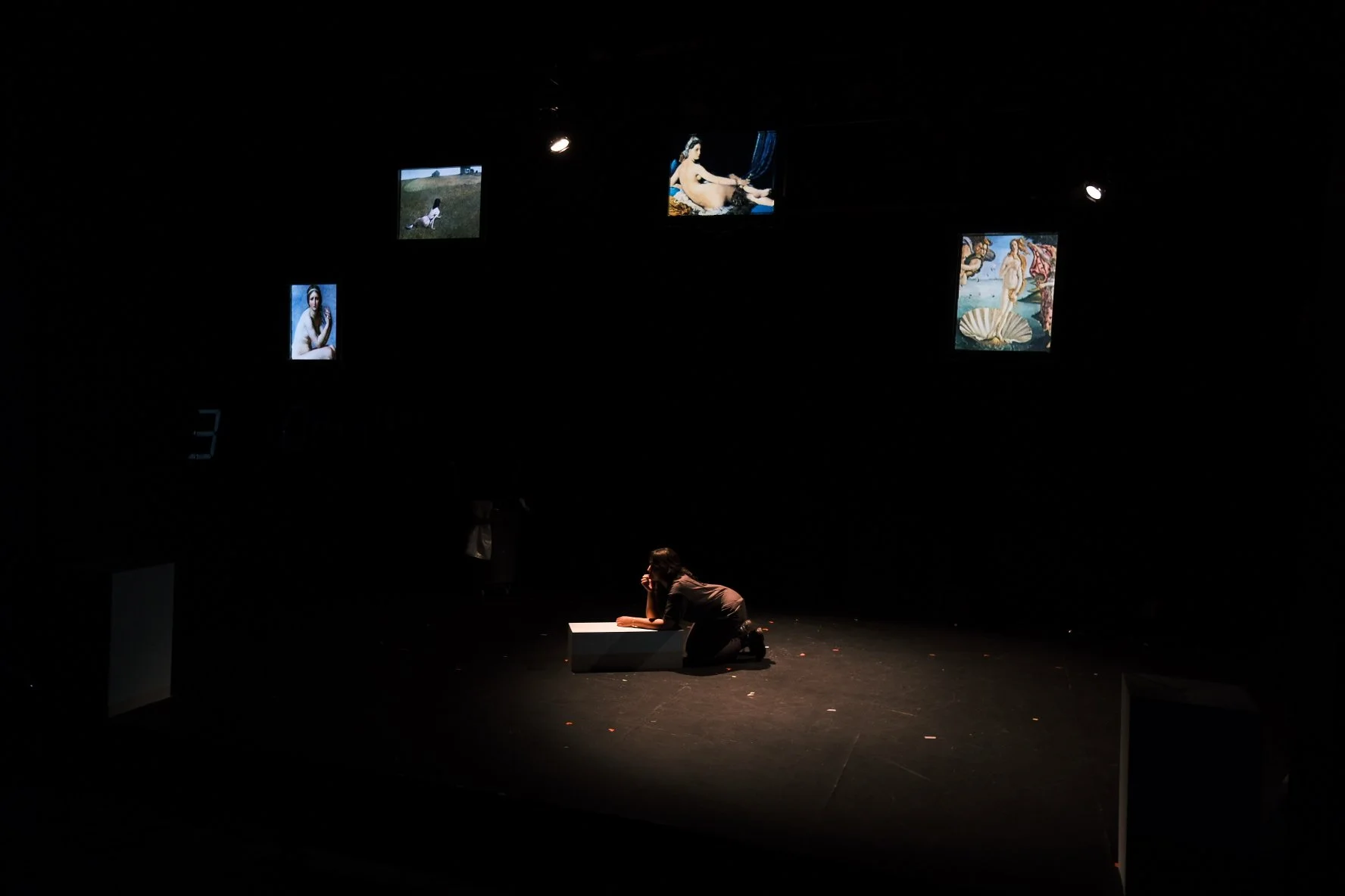 A person kneeling on the stage floor, leaning on a white bench, looking thoughtfully at art images displayed on screens above.