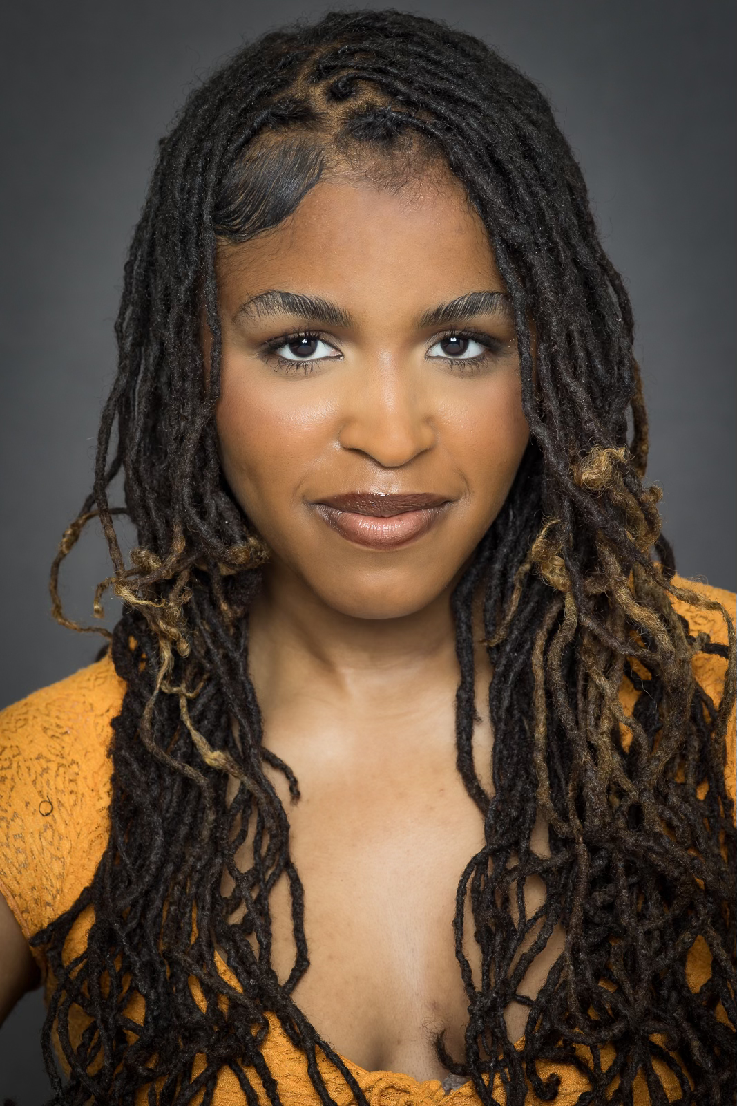 A woman with long dreadlocks and brown skin, smiling slightly, wearing a mustard-colored top against a dark gray background.