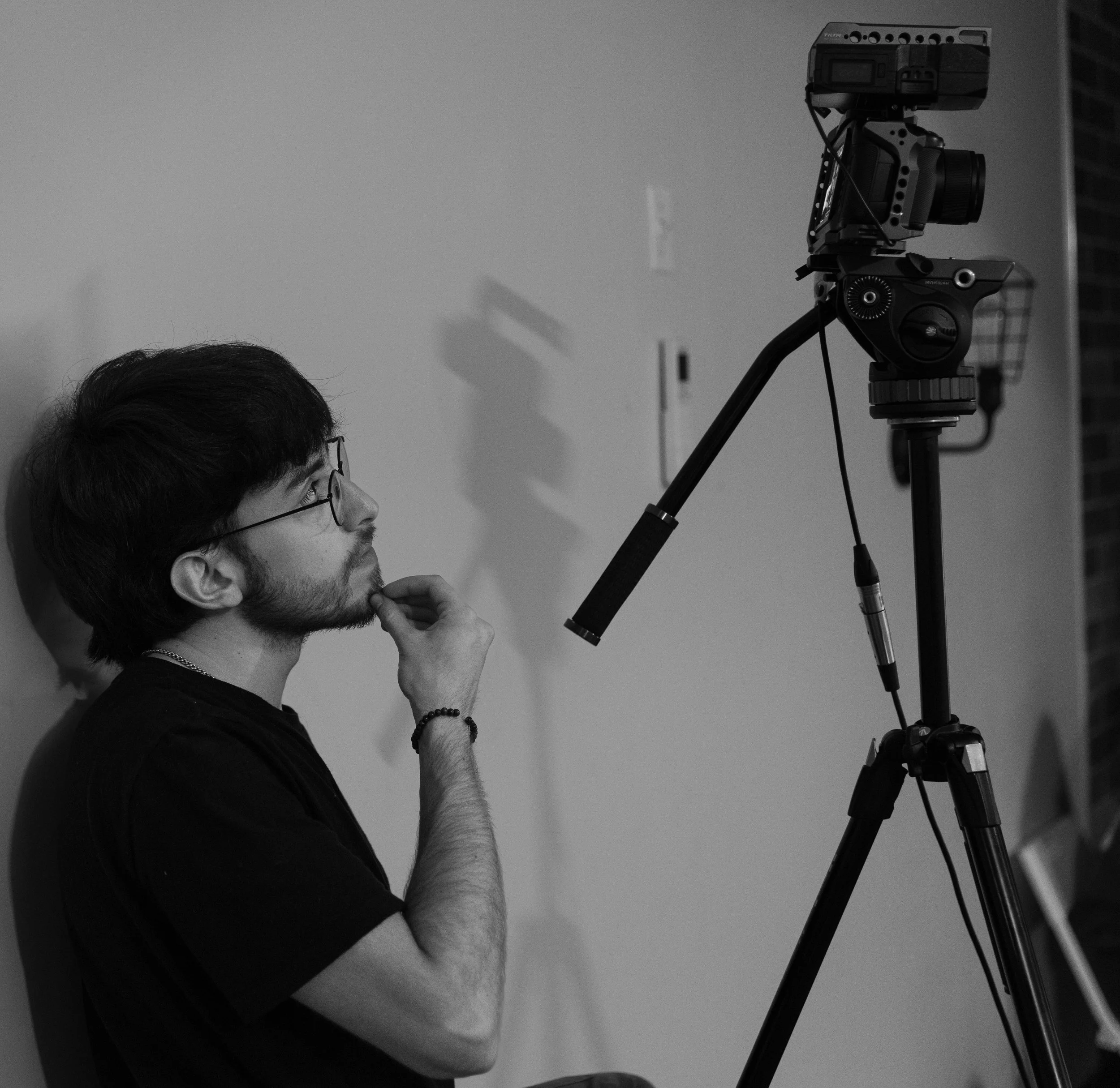 A person with glasses and dark hair is looking through a telescope mounted on a tripod, with their hand resting near their chin.