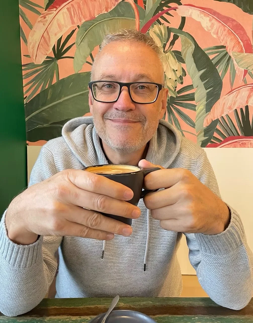 Image of Gary sitting with a coffee at a table and smiling at the camera