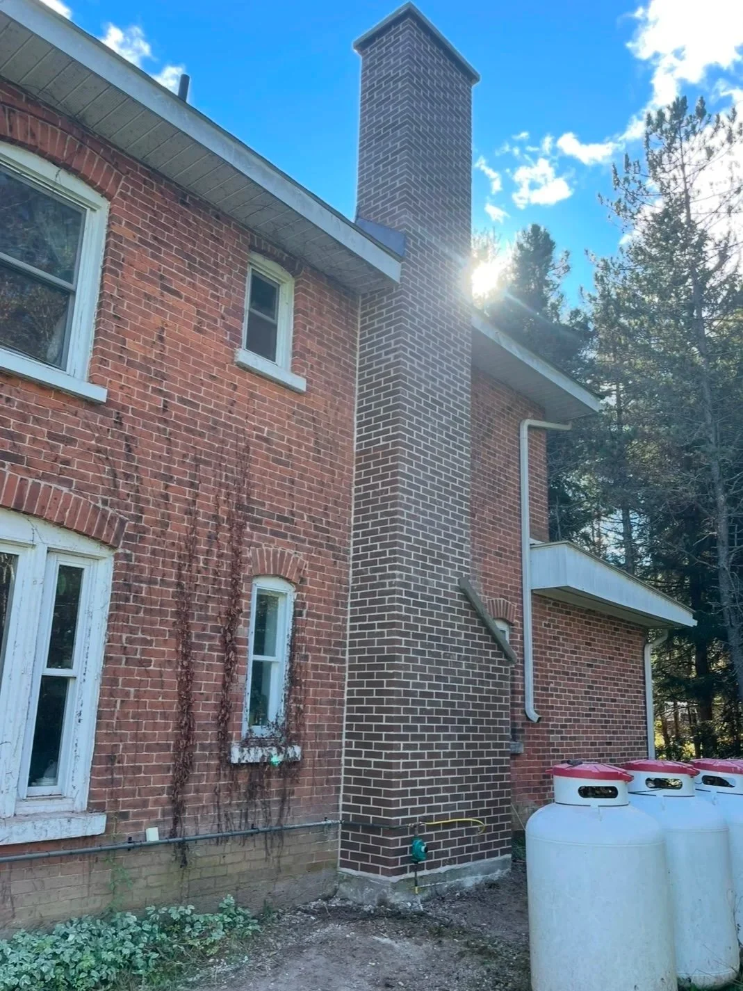 Side of a brick house with small windows, a brick chimney, and propane tanks in the yard, under a partly cloudy sky with sunlight peeking through trees.