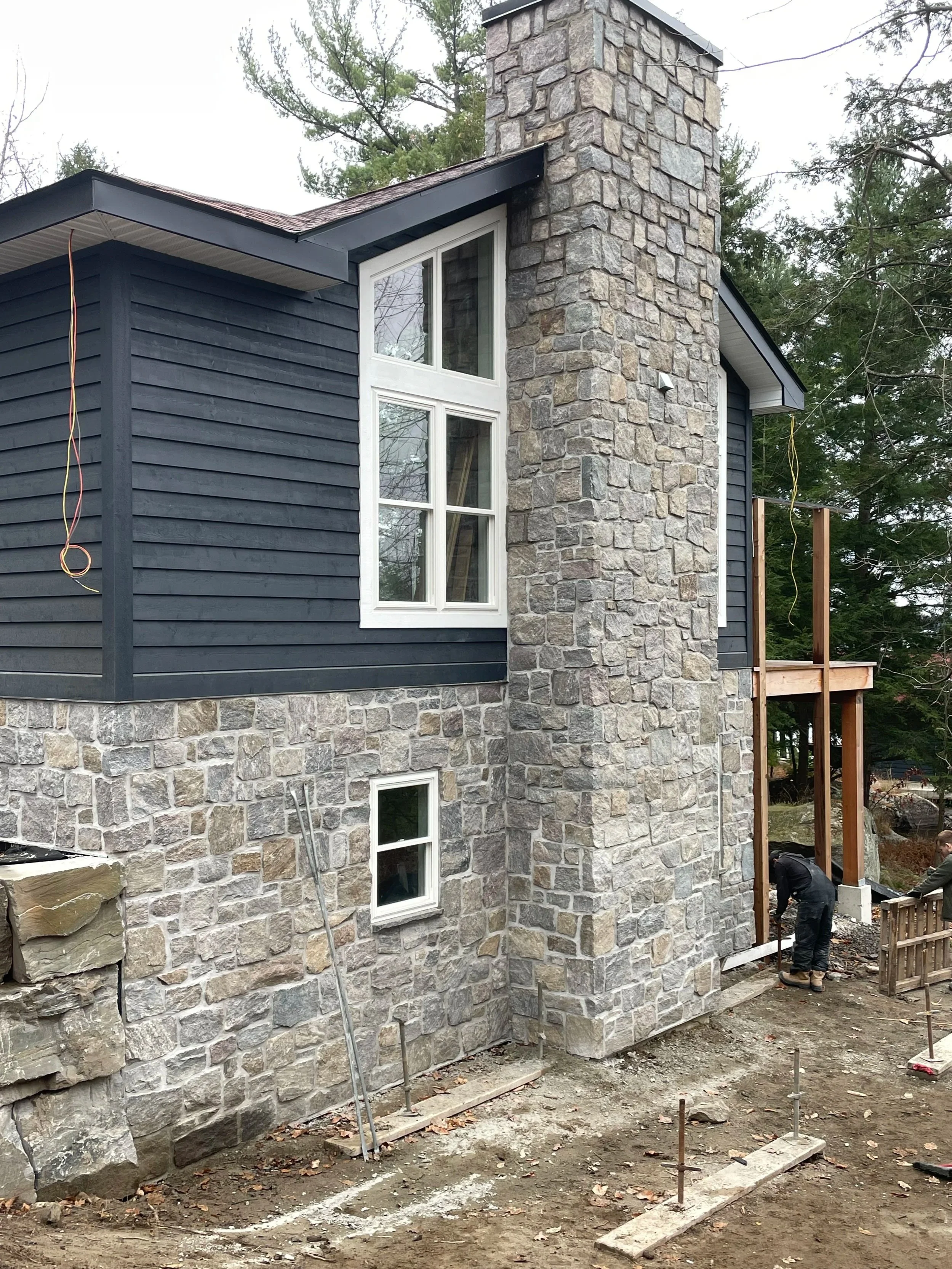 Construction workers building a stone and siding house, with the main structure mostly complete. The house features a large stone chimney, dark gray siding, and a window with white framing. The work area has dirt and construction materials, with wooden posts and workers involved in construction activities.