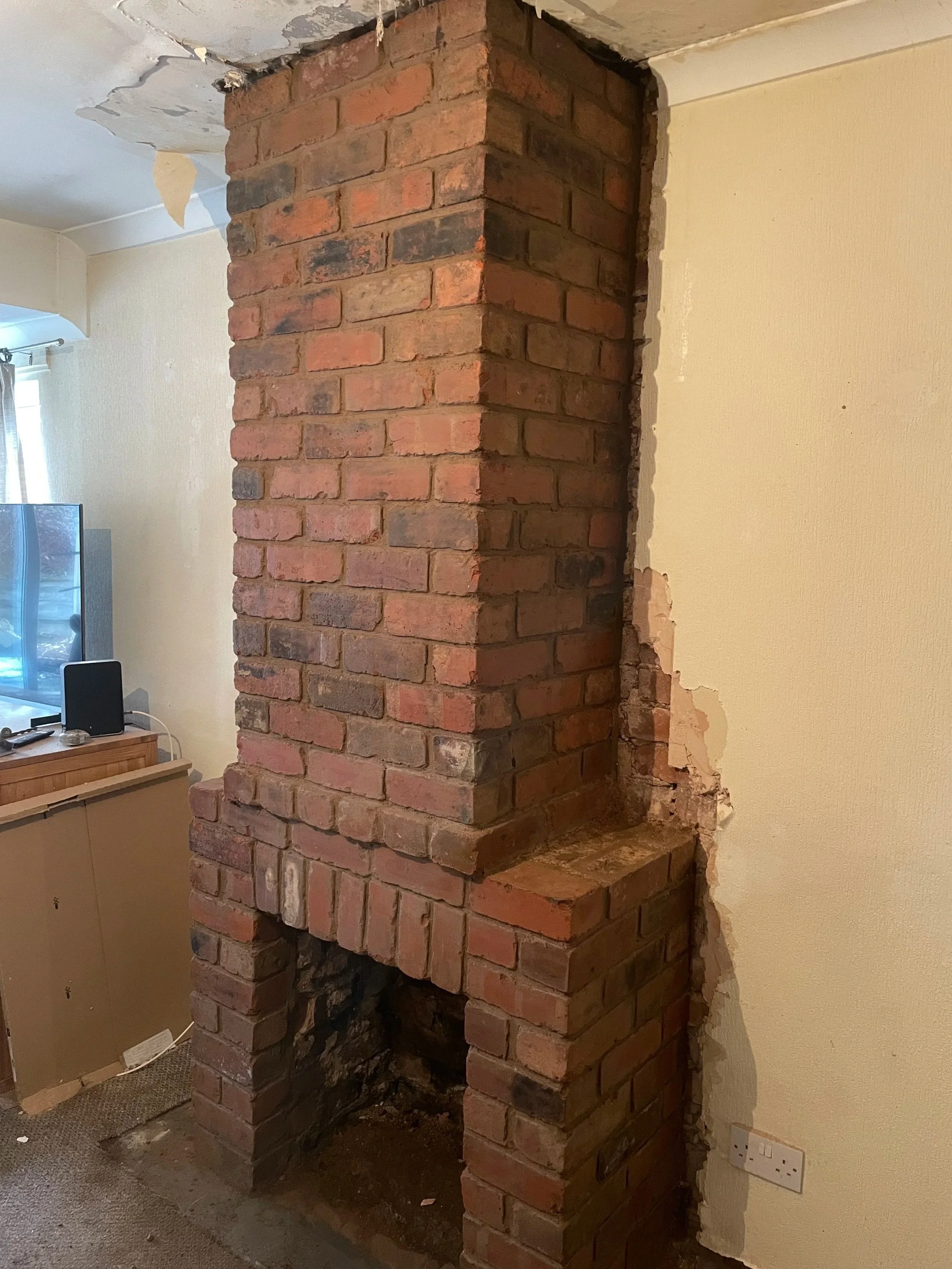 A brick chimney in the process of renovation inside a home. The bricks are red with blackened and weathered areas. The chimney is adjacent to a cream-colored wall and the ceiling shows signs of damage with peeling paint.