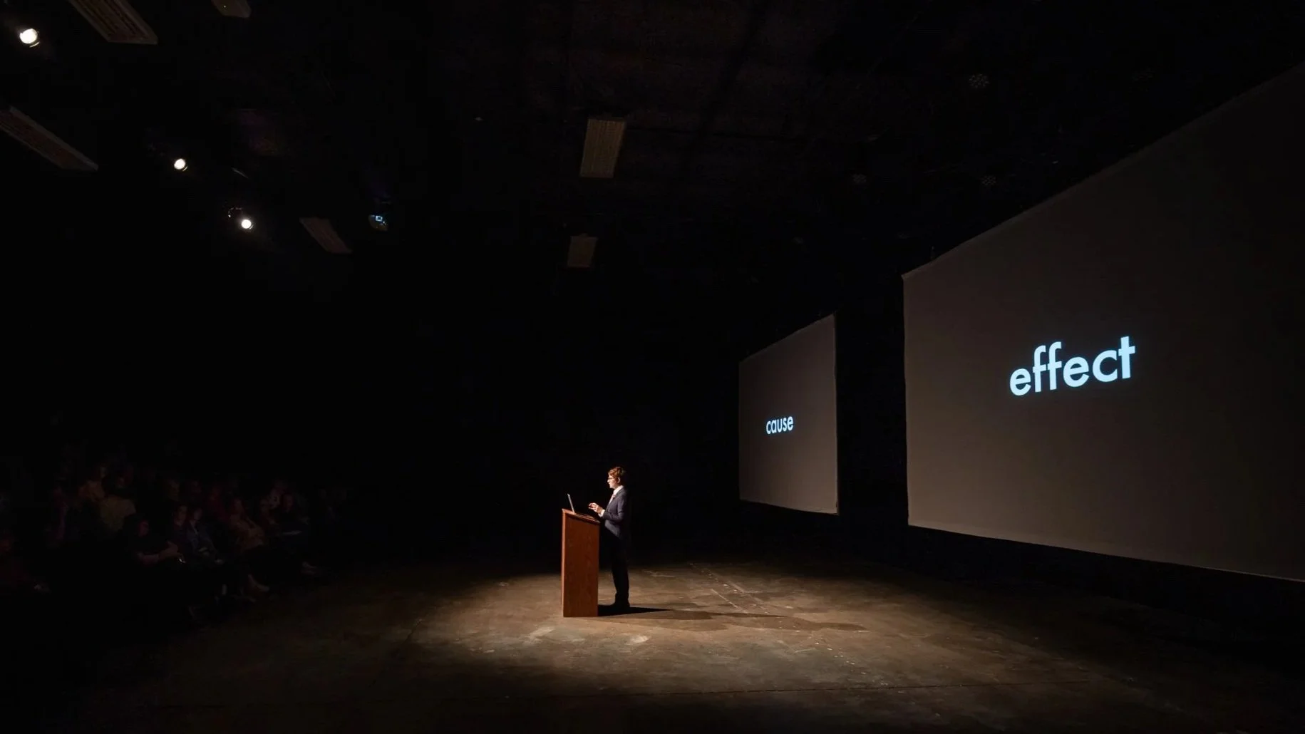 Bean Gilsdorf lectures at a podium. Behind her are two massive screens that read "cause" and "effect."