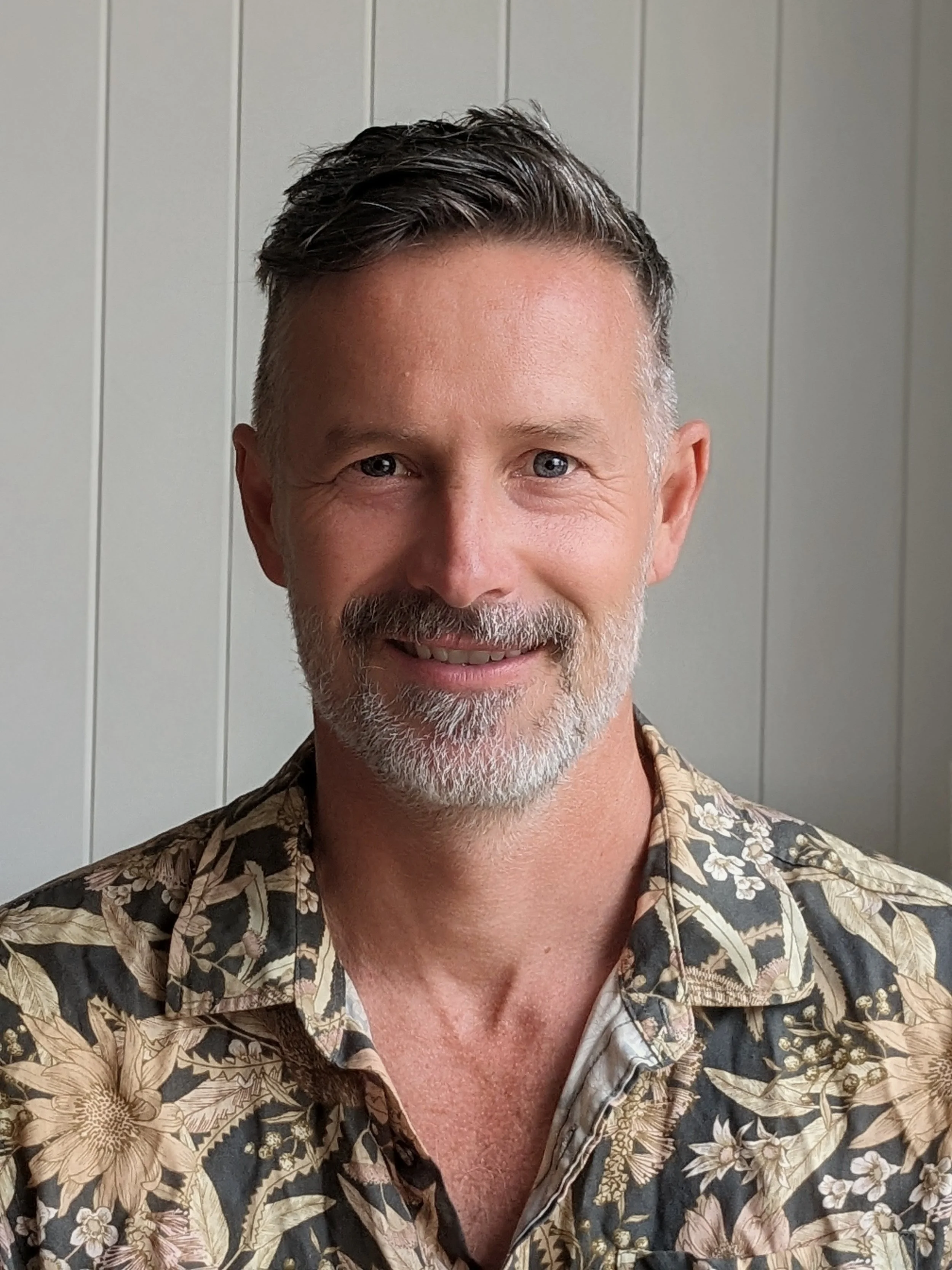 A middle-aged man with short, dark, and gray hair, and a beard, smiling while wearing a patterned shirt with floral designs, standing in front of light gray vertical paneling.