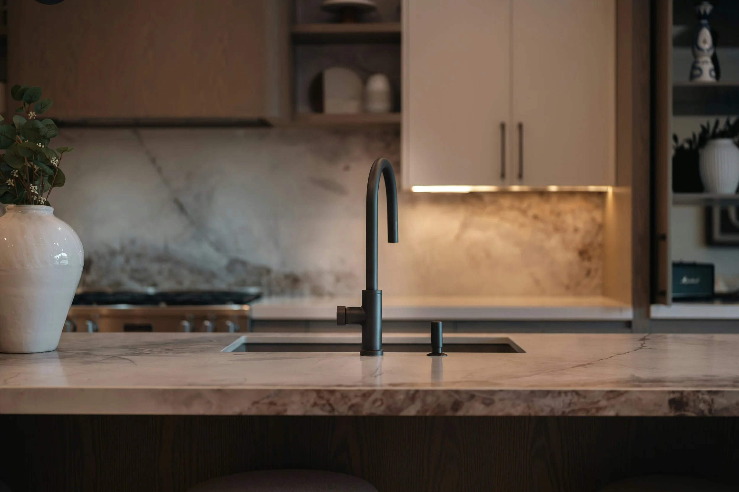 A modern kitchen with a black faucet over a white marble countertop, with built-in cabinets and decorative plants in the background.