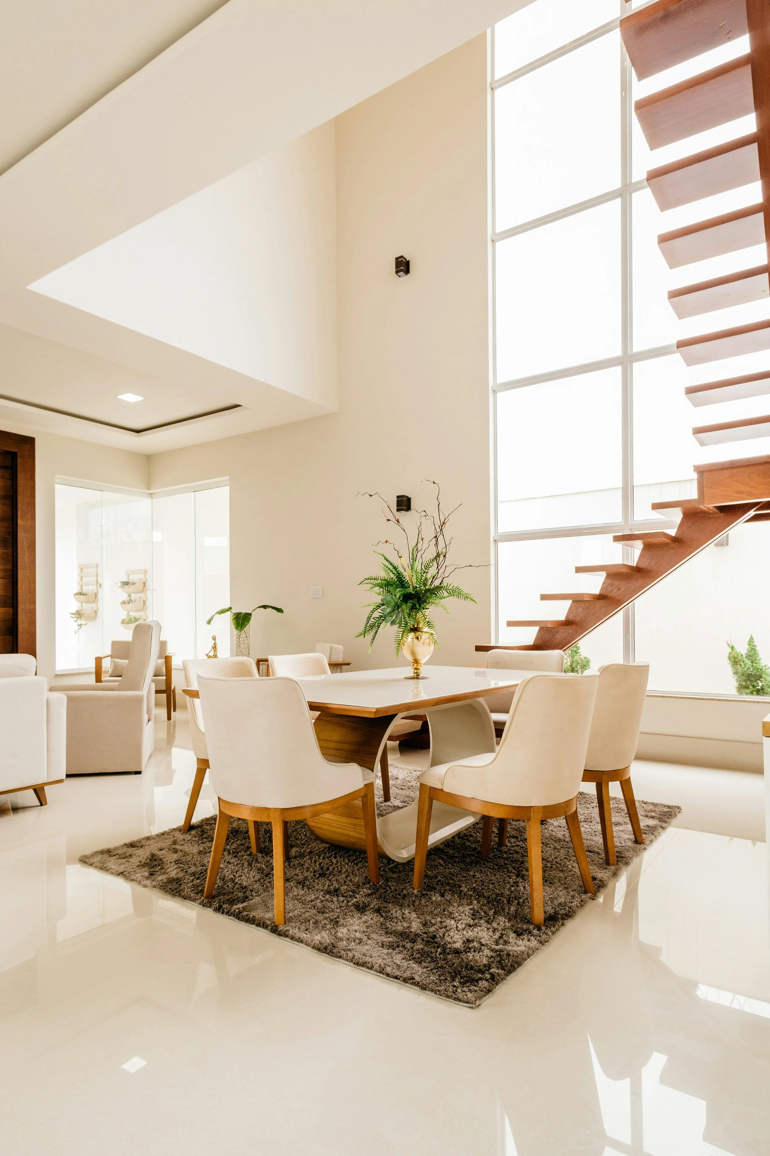 Bright, modern dining room with a large wooden table, cream chairs, a gray rug, large windows, and a tall staircase along the wall with wooden steps.