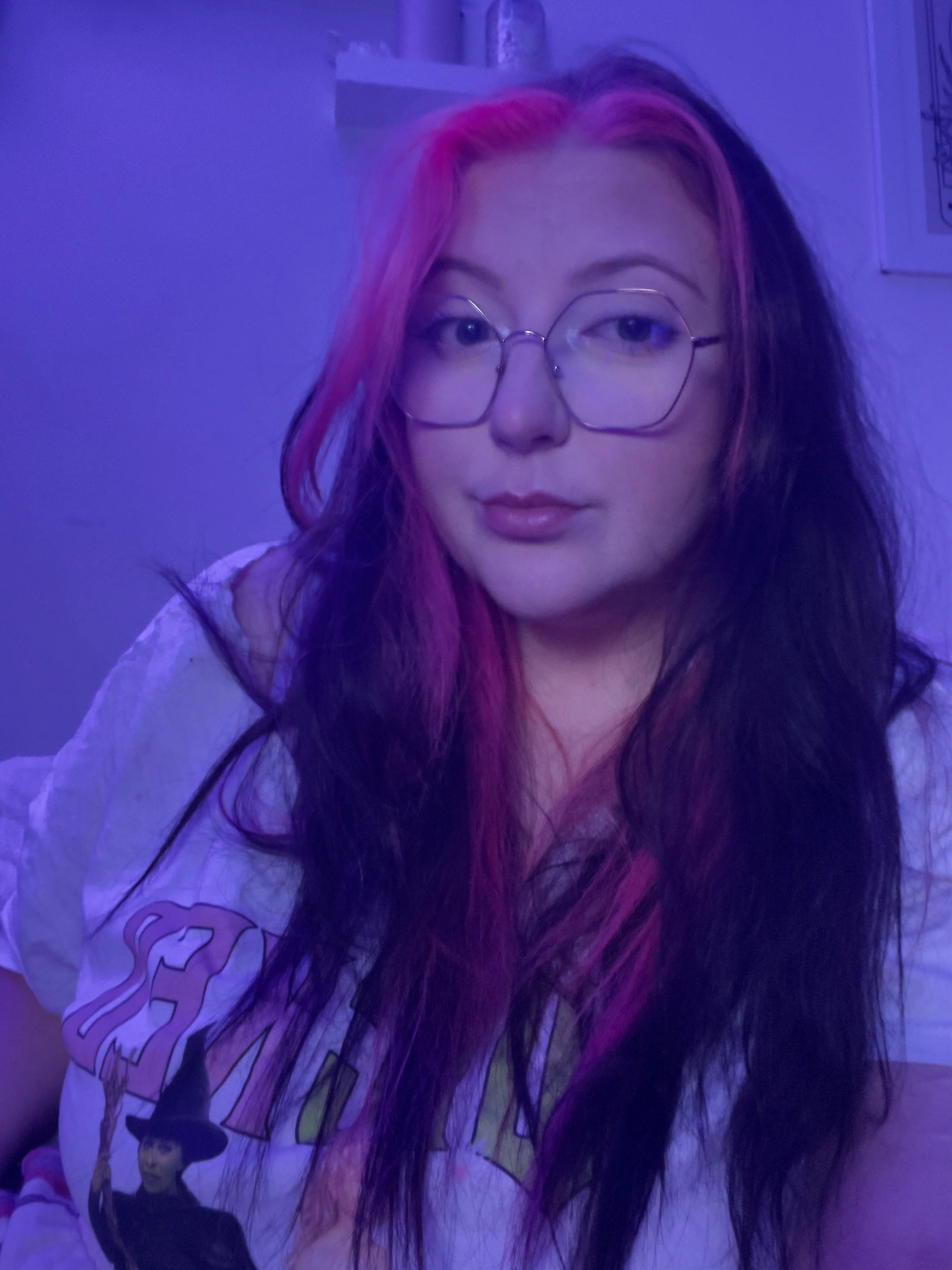 A woman with long dark hair with pink highlights and wearing glasses, illuminated by purple lighting, taking a selfie in a room with posters on the wall.