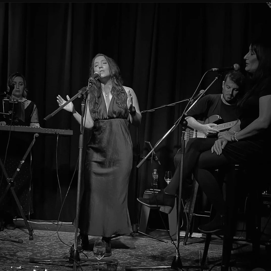 Black and white photo of a woman singing into a microphone on stage, accompanied by a guitarist and other musicians, with dark curtains in the background.