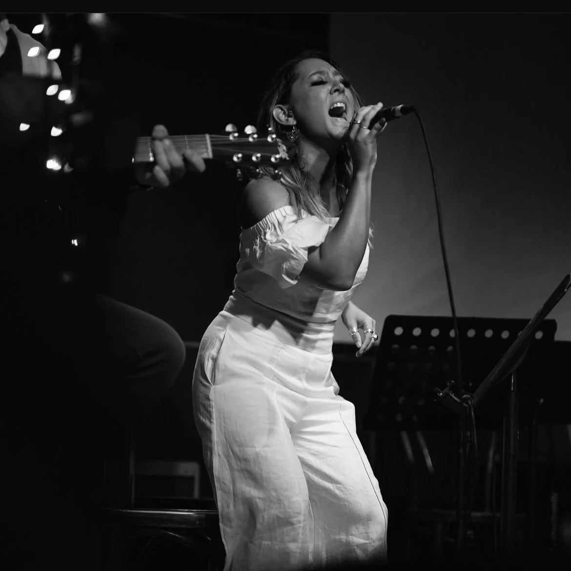 A woman singing passionately into a microphone while a person plays guitar beside her in a dark setting.