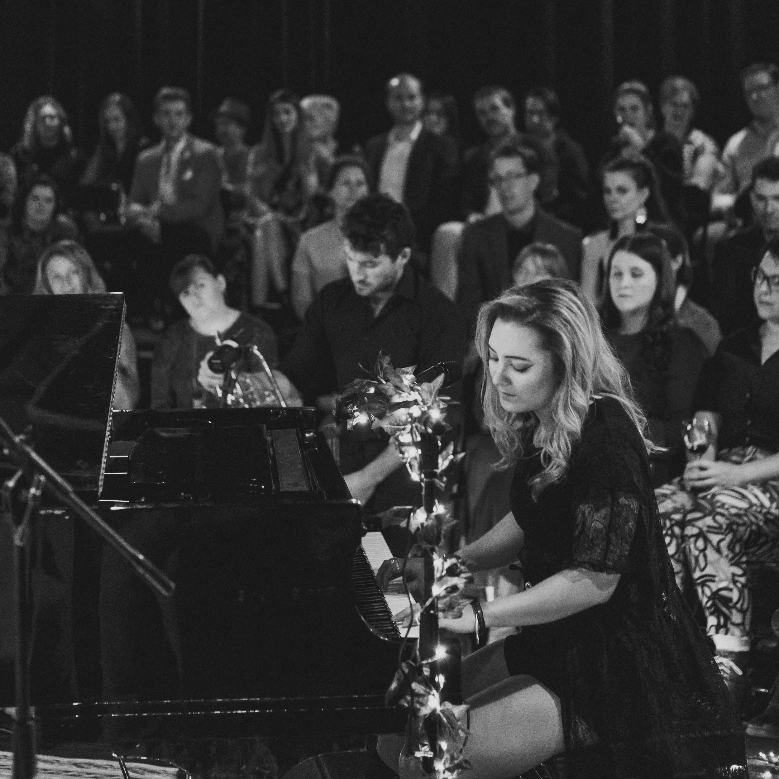 People gathered at a show, some standing and some sitting around a grand piano, as Alexis Naylor performs.
