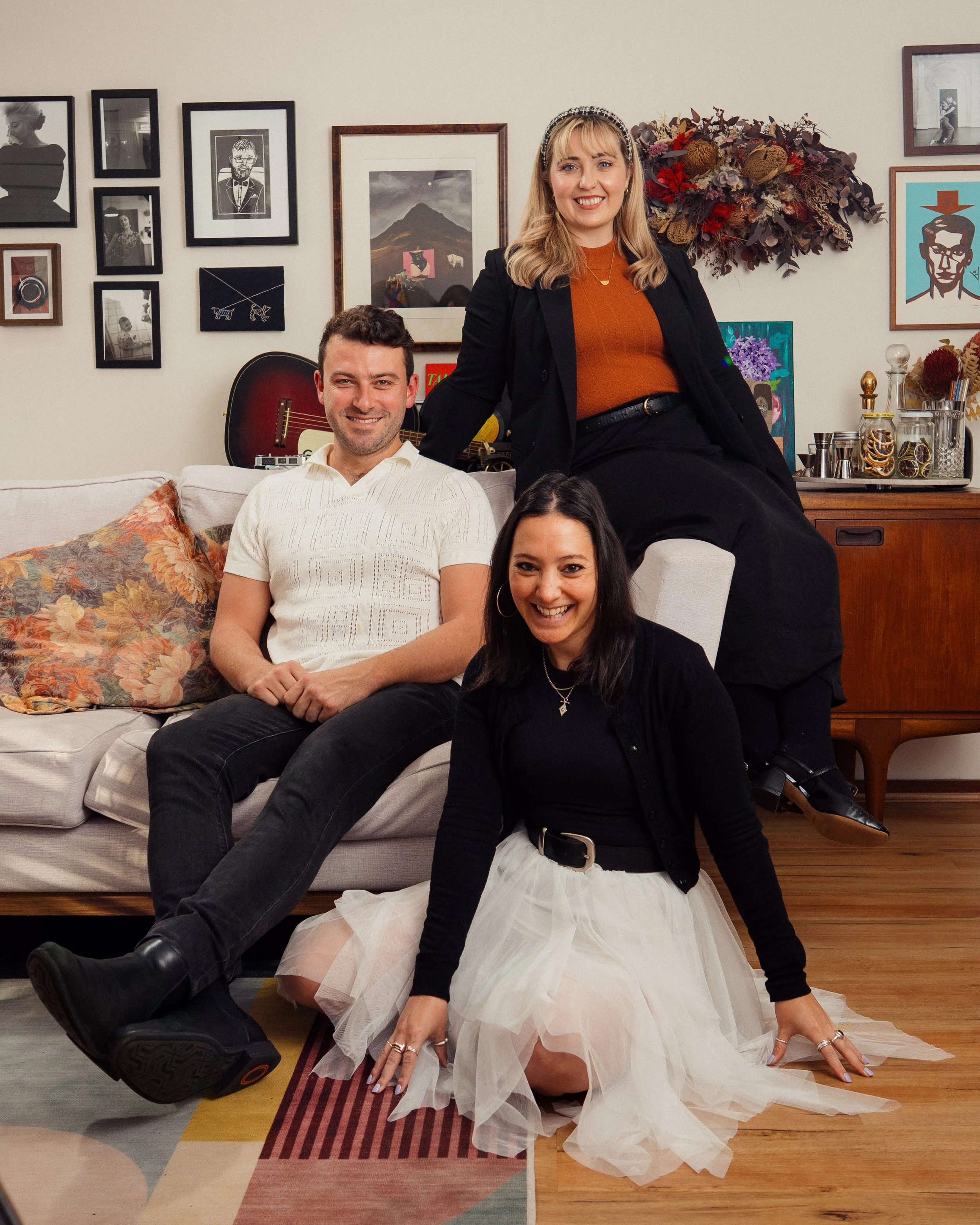 Group of four people in a living room, with a woman in a black blazer sitting on a couch, a man in a white shirt sitting next to her, and a woman in a black top and white skirt kneeling on the floor, while another woman in black is partially visible. The background has framed photos, artwork, and decorative items.