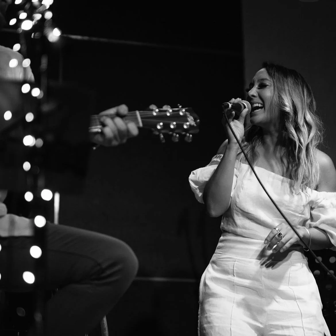 Alexis Naylor singing into a microphone while a musician Mark Turner guitar beside her, in a black-and-white photo.