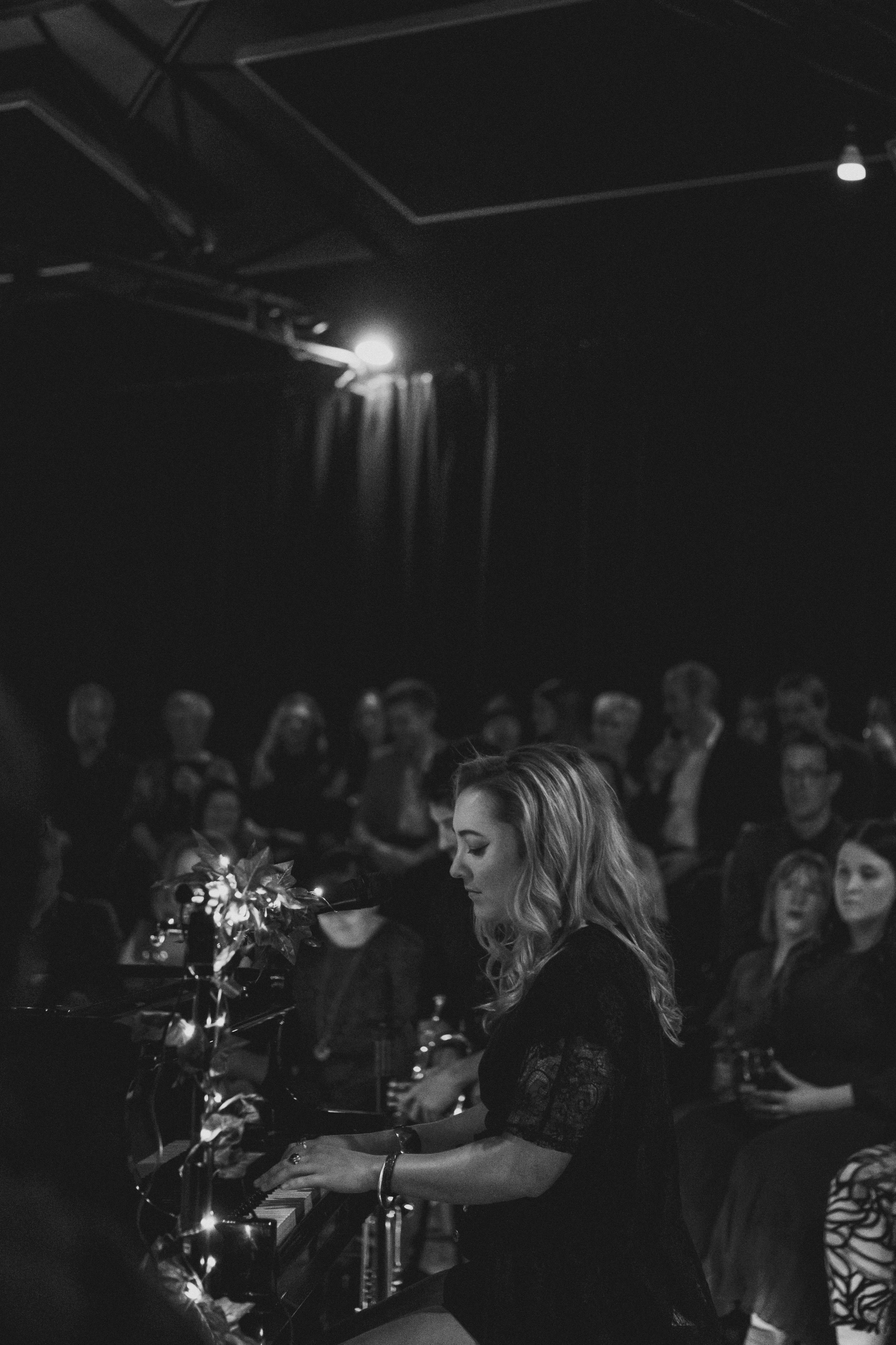 Alexis Naylor playing the piano at a crowded indoor event with many people in the background, some standing and watching, in a dimly lit space.