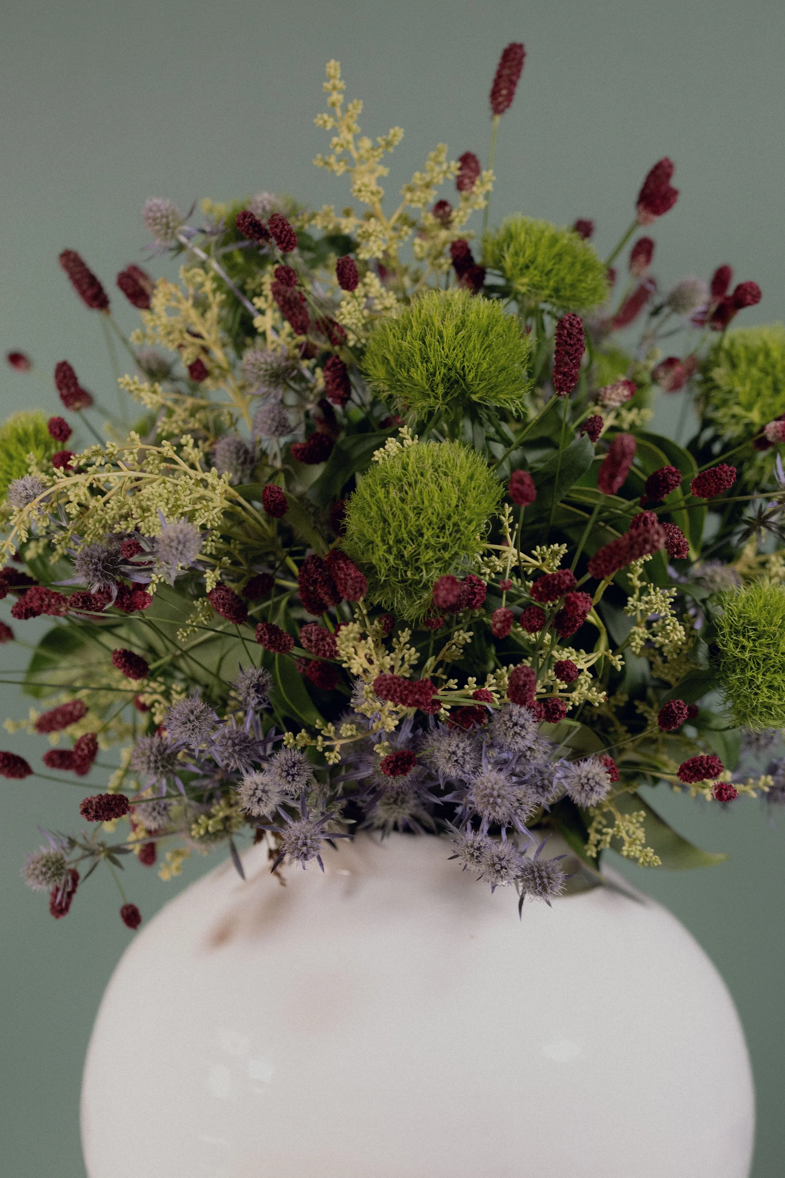 Un bouquet di fiori vari di colore, disposto in un vaso bianco con forma rotonda, su uno sfondo verde chiaro.