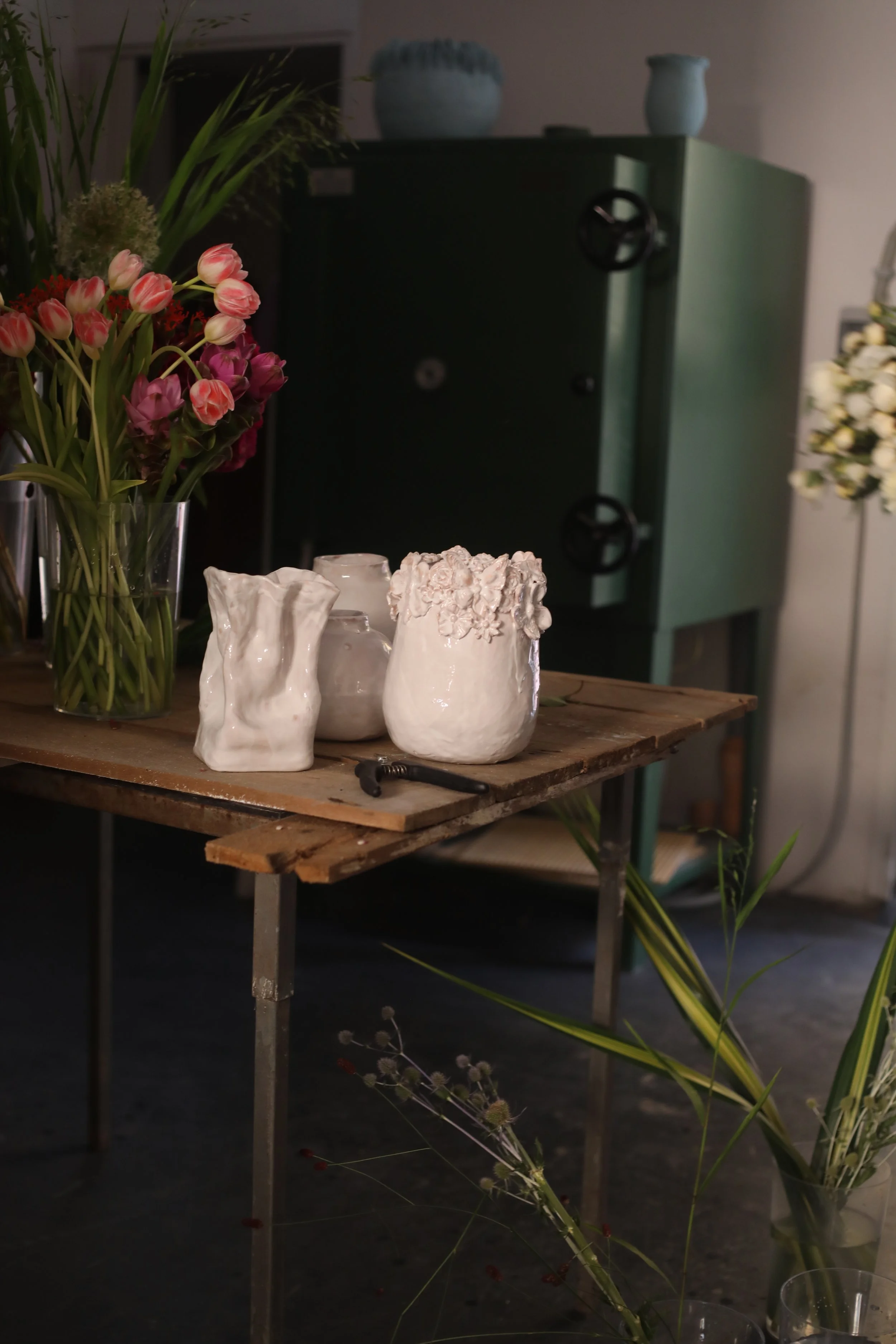 Un tavolo di legno con vasi di ceramica bianchi decorati con fiori e un mazzo di fiori in un grande vaso di vetro, con un armadio verde scuro sullo sfondo.