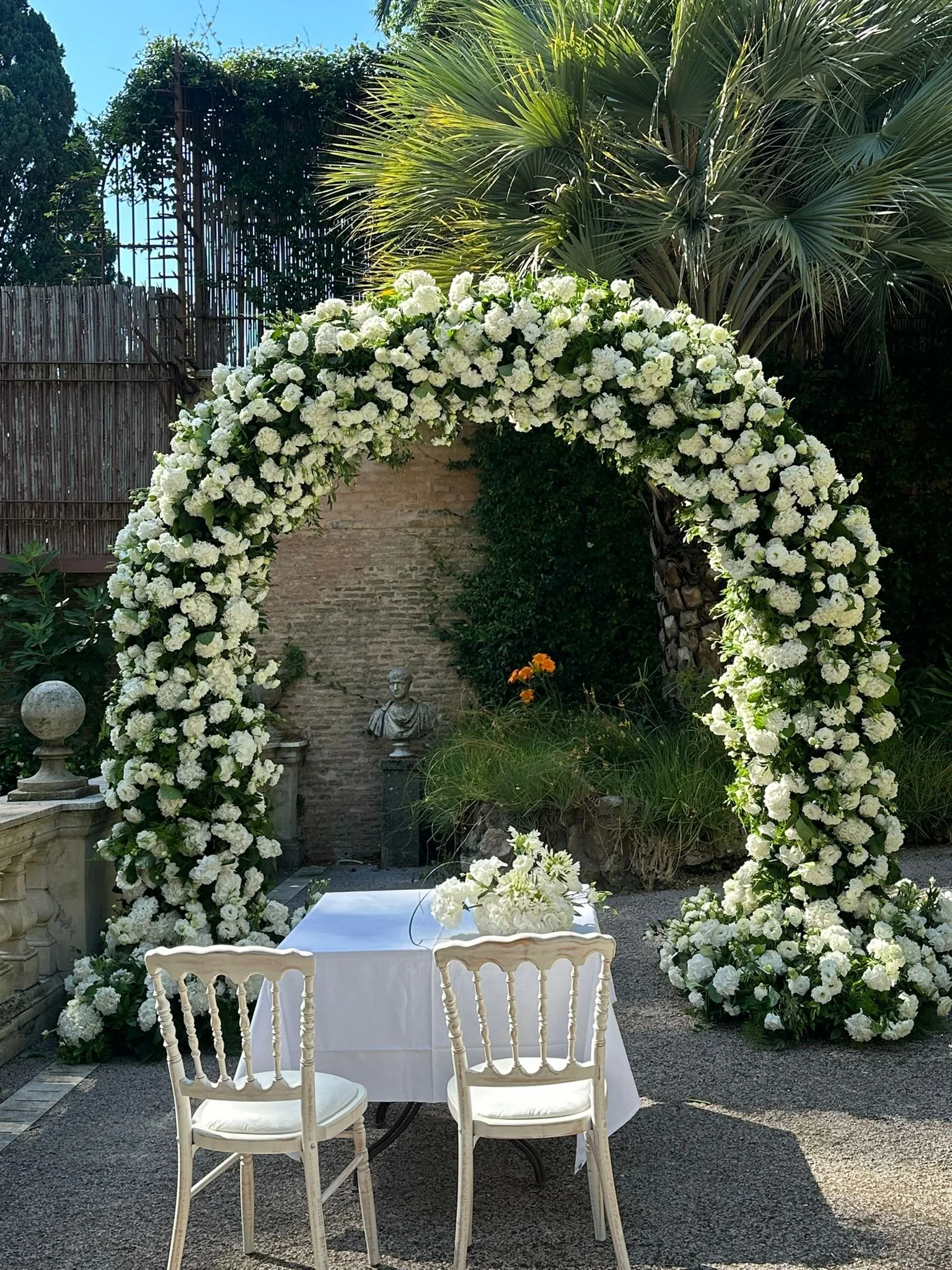 Un arco decorato con fiori bianchi, un tavolo con una tovaglia bianca e sedie in legno, in un giardino con statue, piante e un muro di mattoni, ambiente per un matrimonio o evento.