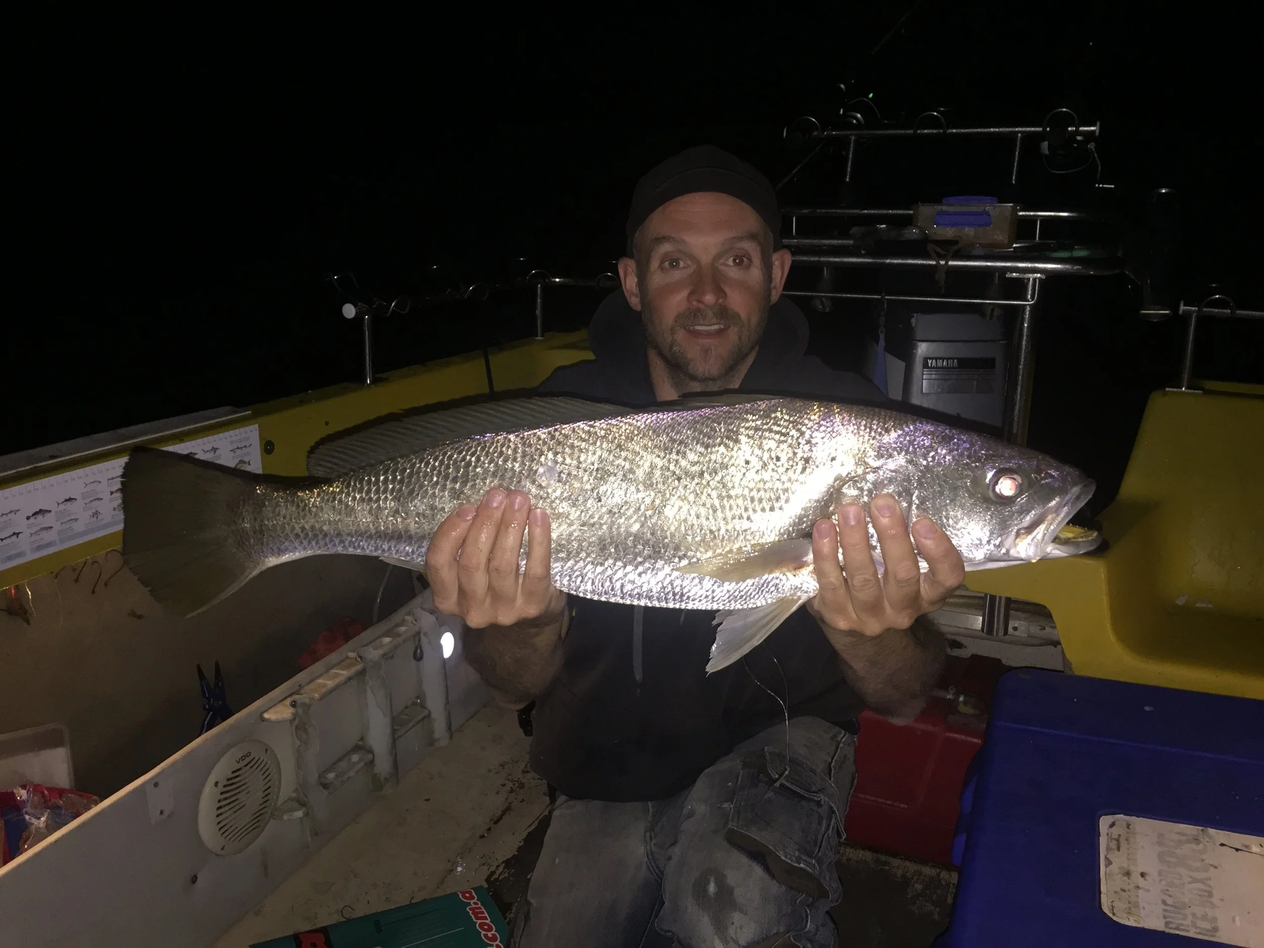 A man holding a large fish on a boat at night with fishing gear in the background.