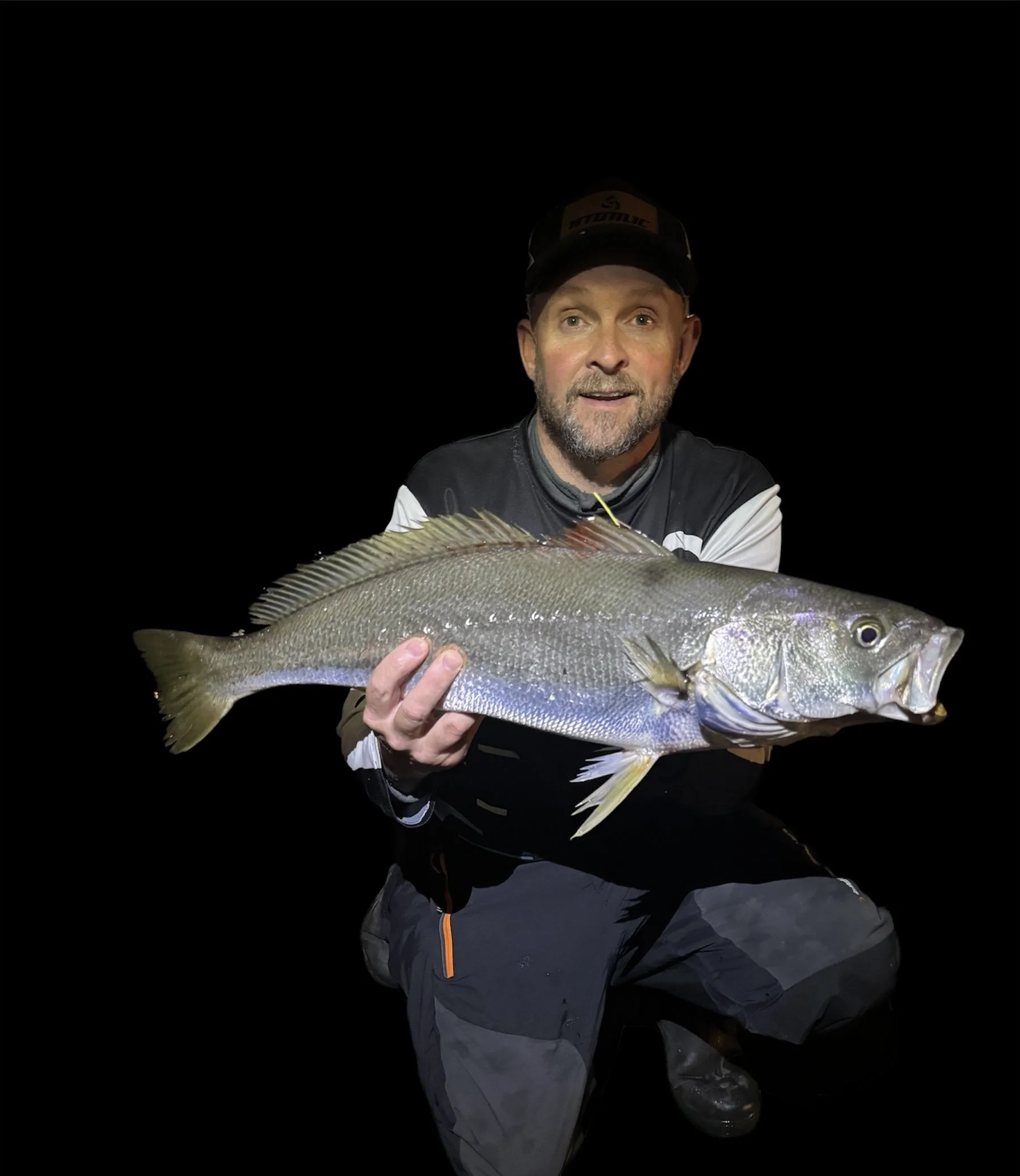 A man in dark clothing and a black cap holding a large fish against a black background at night.