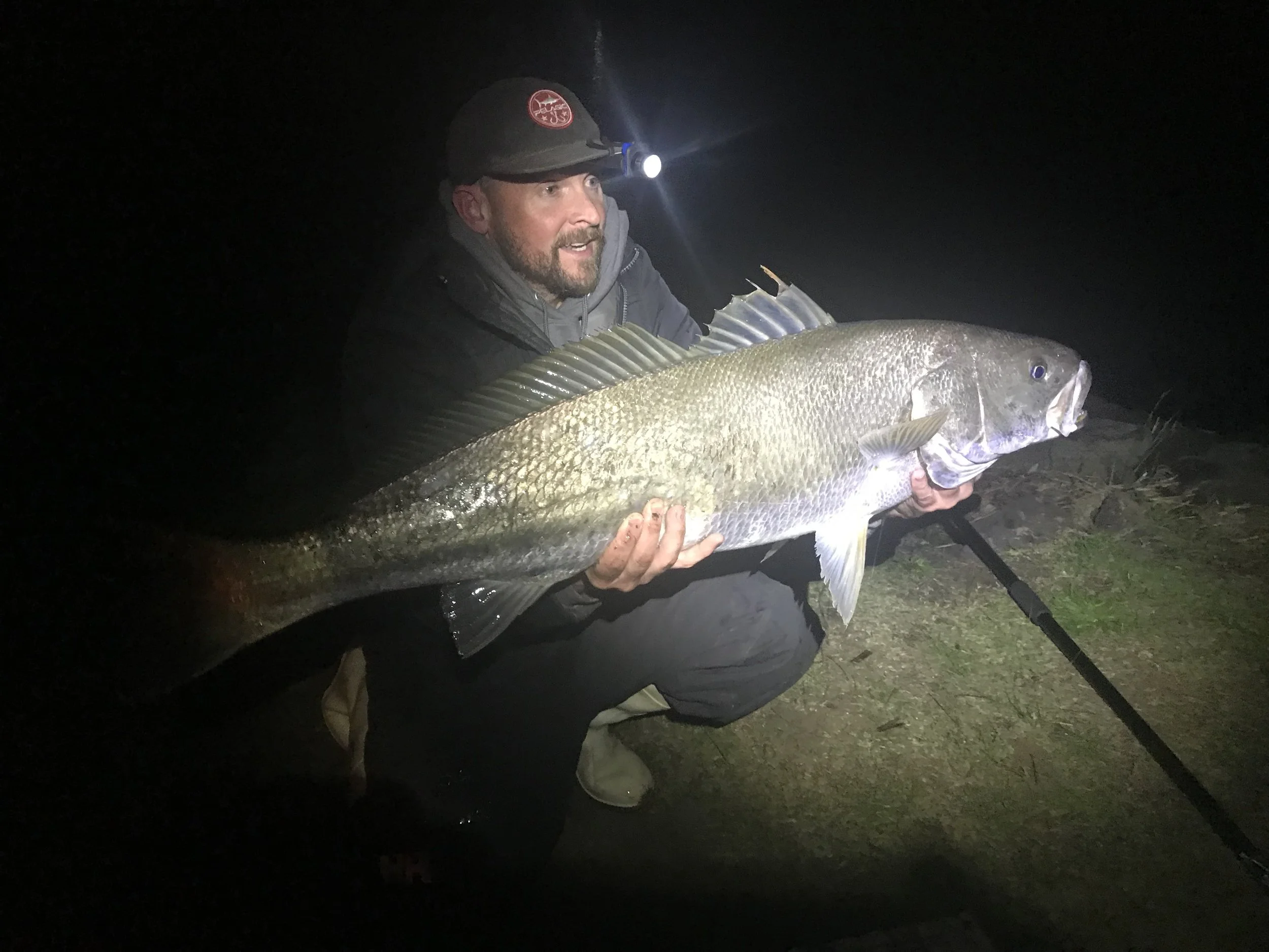 A man in a dark jacket and cap with a headlamp, holding a large fish at night outdoors.