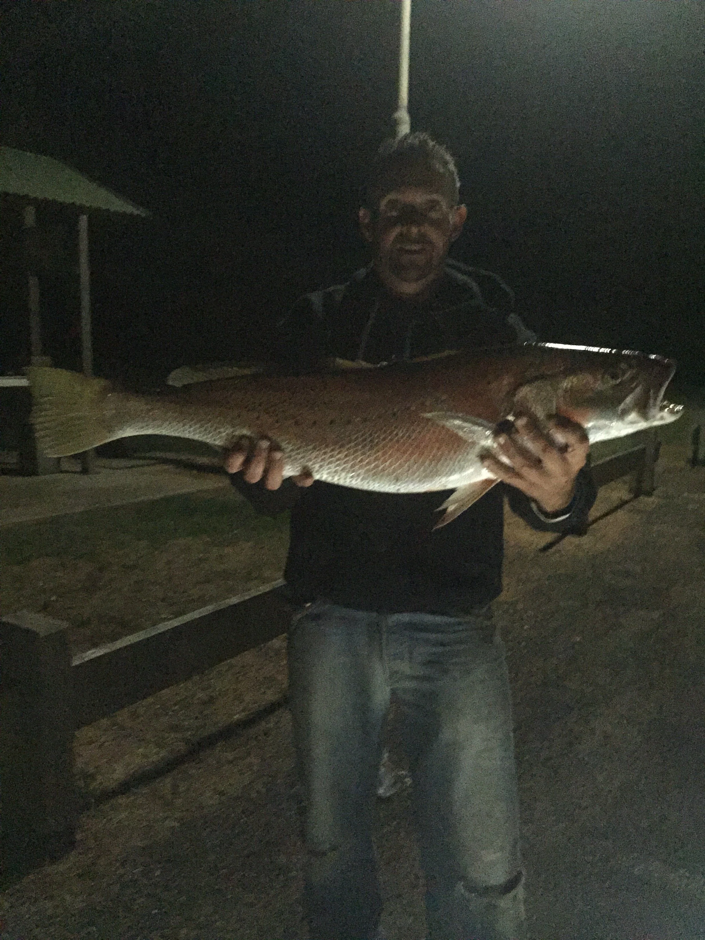 A person is holding a large fish, standing outdoors at night near a bench and an umbrella.