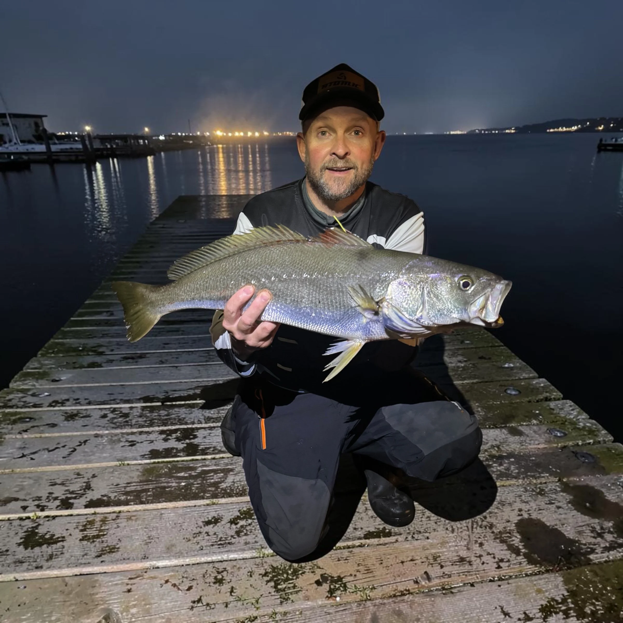 A man with a beard wearing a cap and a dark jacket, kneeling on a dock at night, holding a large fish. The background shows a calm body of water with boats, docks, and distant lights under a dark sky.