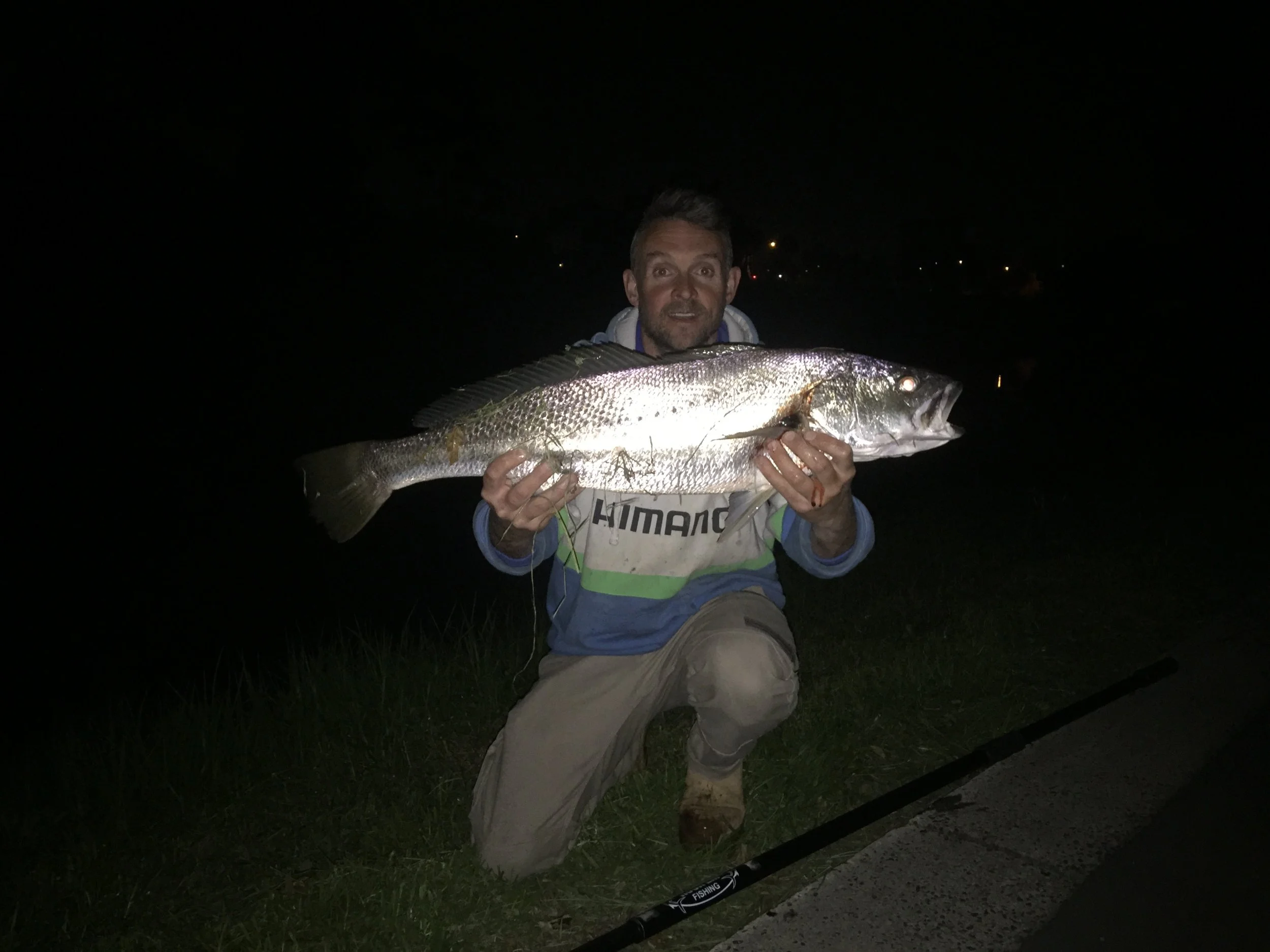 A man kneeling on the grass at night holding a large fish. The background is dark with a few distant lights. The man is wearing outdoor clothing, and a fishing rod is on the ground nearby.