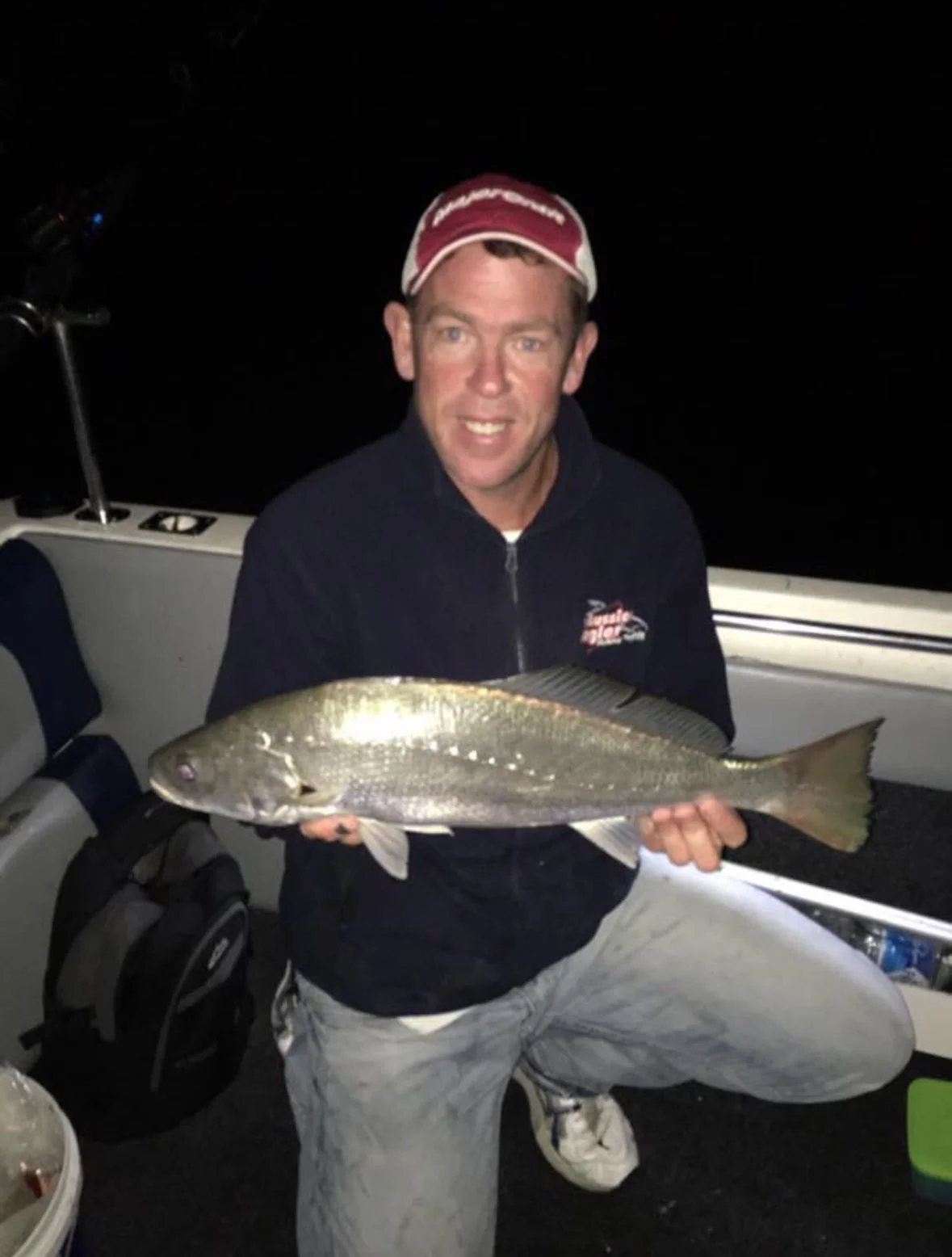 A man wearing a red and white cap and a navy jacket holding a large fish on a boat at night.