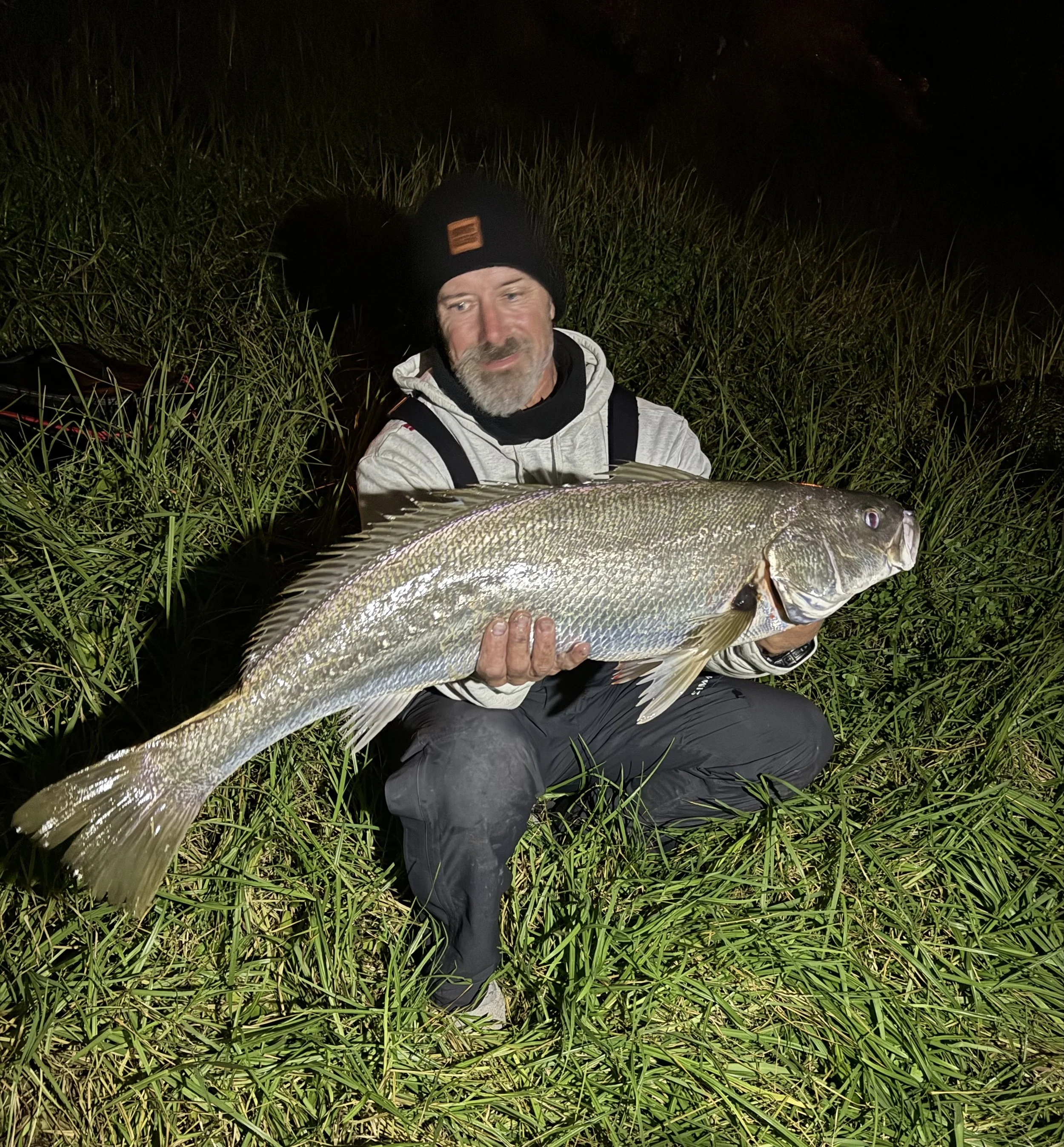 Man in a black beanie, gray jacket, and black pants kneeling in tall grass at night, holding a large fish horizontally in front of him.