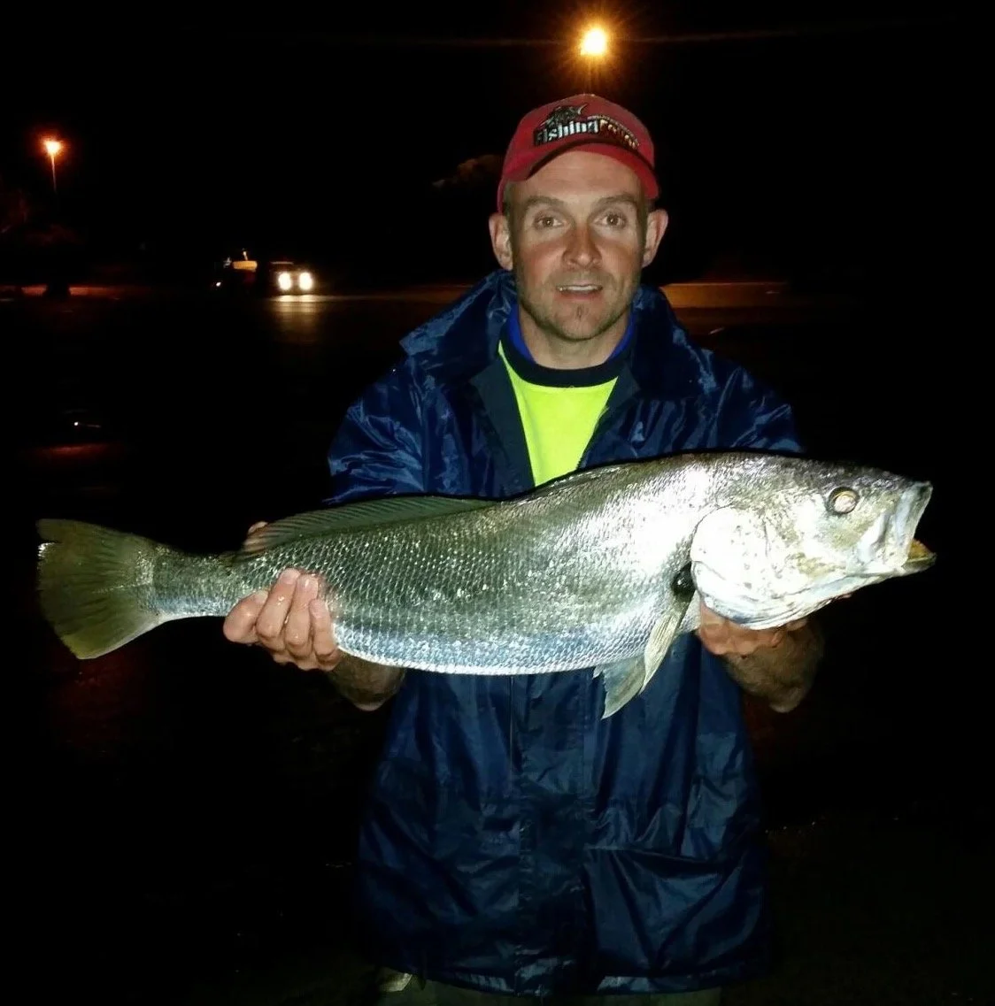A man in a dark rain jacket and red cap holding a large fish at night, illuminated by camera flash with cars and streetlights in the background.