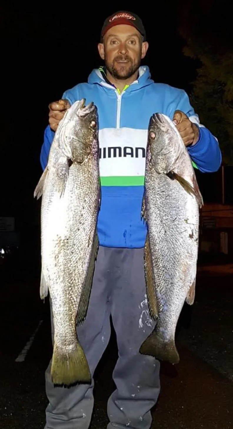 A man wearing a blue and white hoodie and gray pants holding two large fish, one in each hand, outdoors at night.