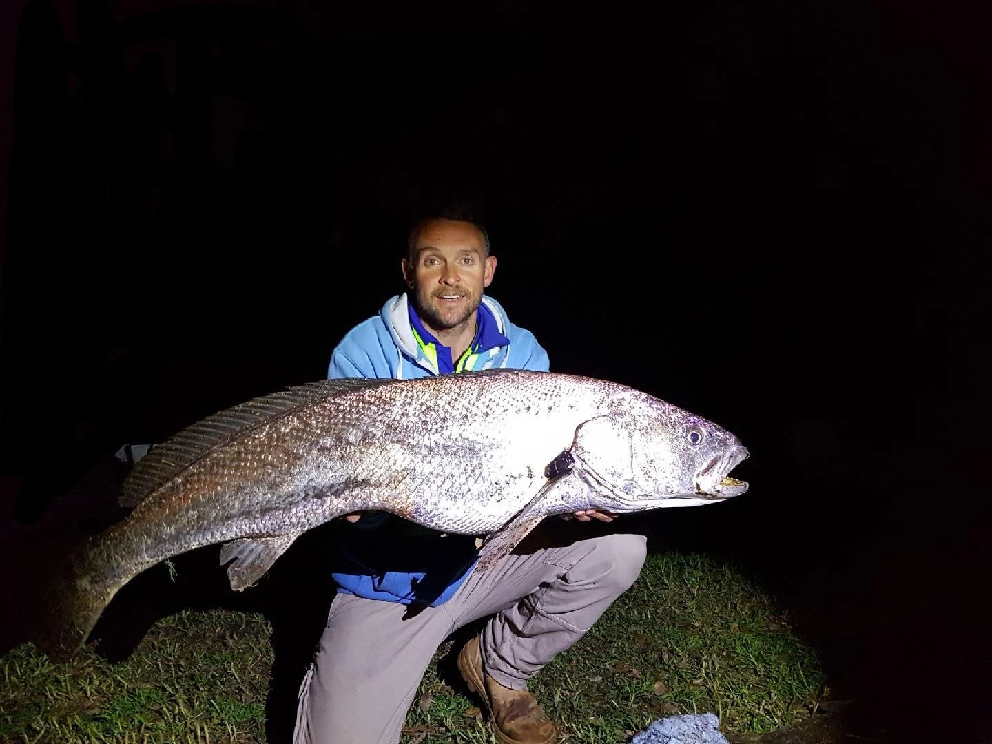 A man kneeling on the grass at night, holding a large fish, with a dark background.