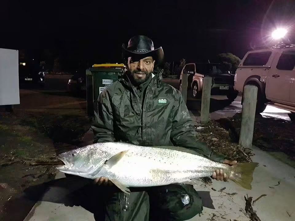 Man wearing a cowboy hat and raincoat holding a large fish in a parking lot at night.