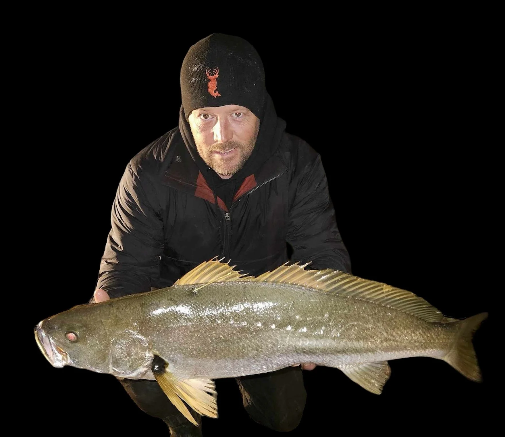 A man holding a large fish, wearing a black beanie with a red lobster logo, and dressed in a black jacket, against a black background.