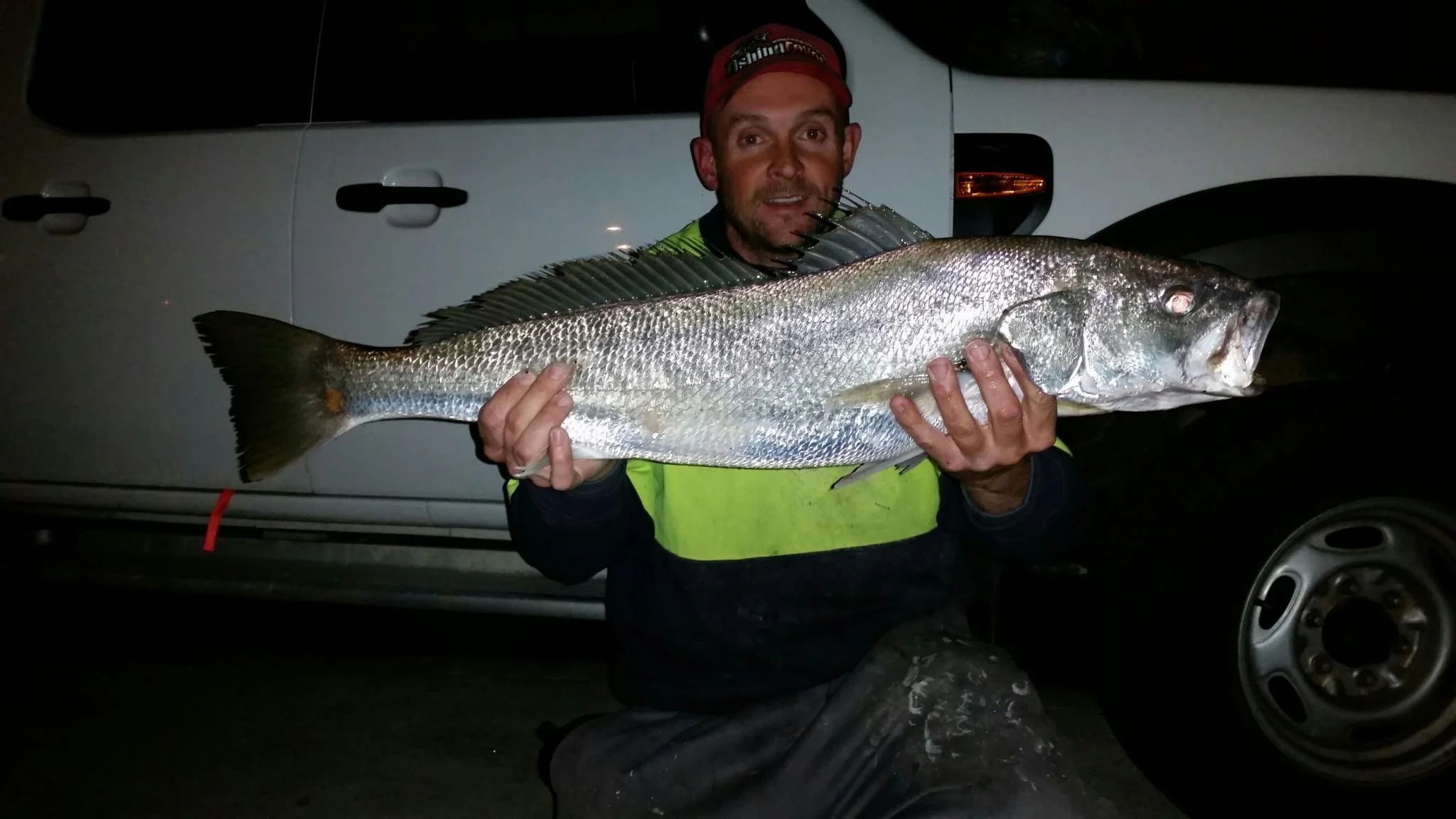 Man holding a large fish at night beside a white vehicle.