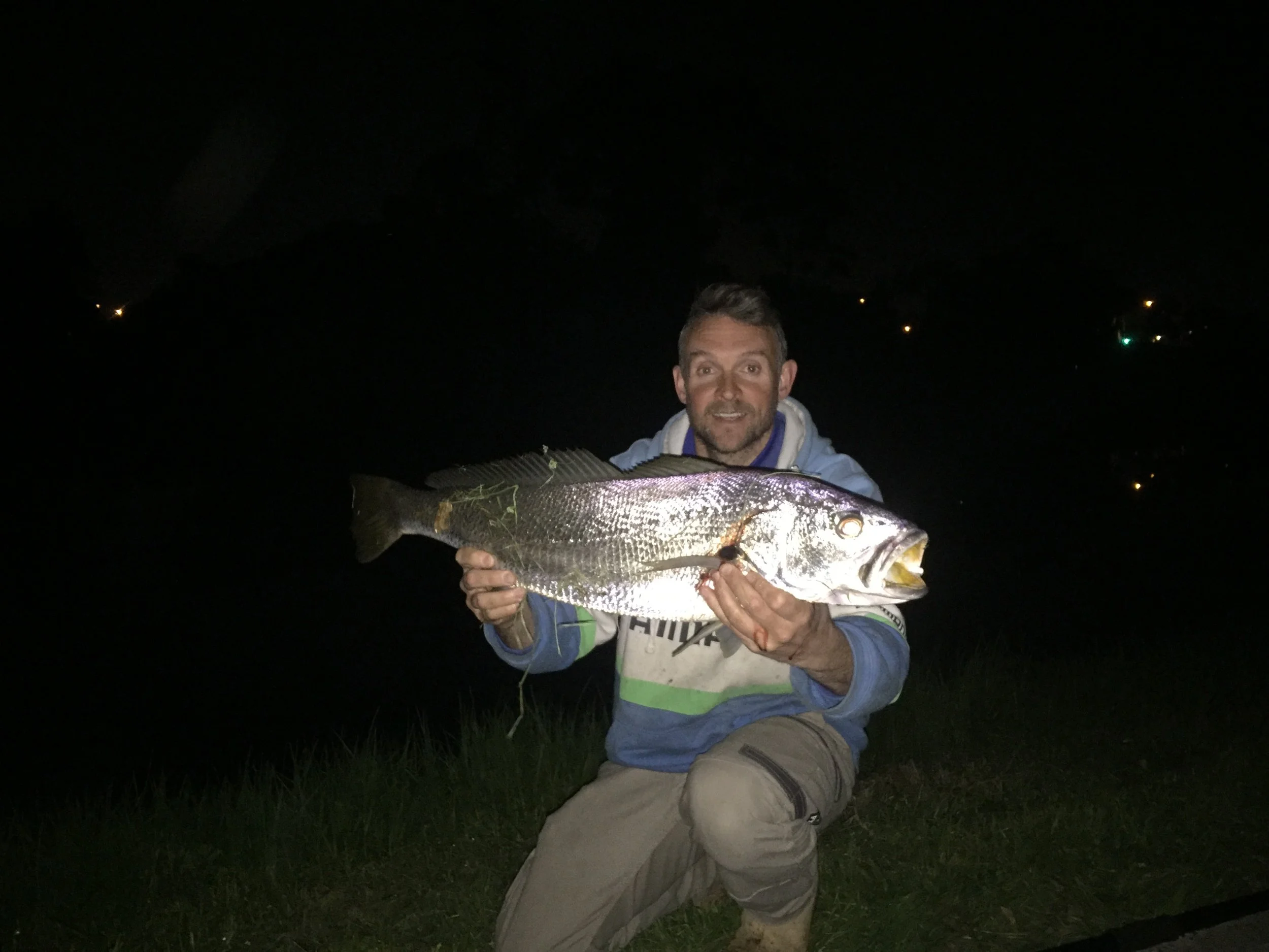 A man kneeling on grass at night, holding a large fish he caught, with a dark sky and distant lights in the background.