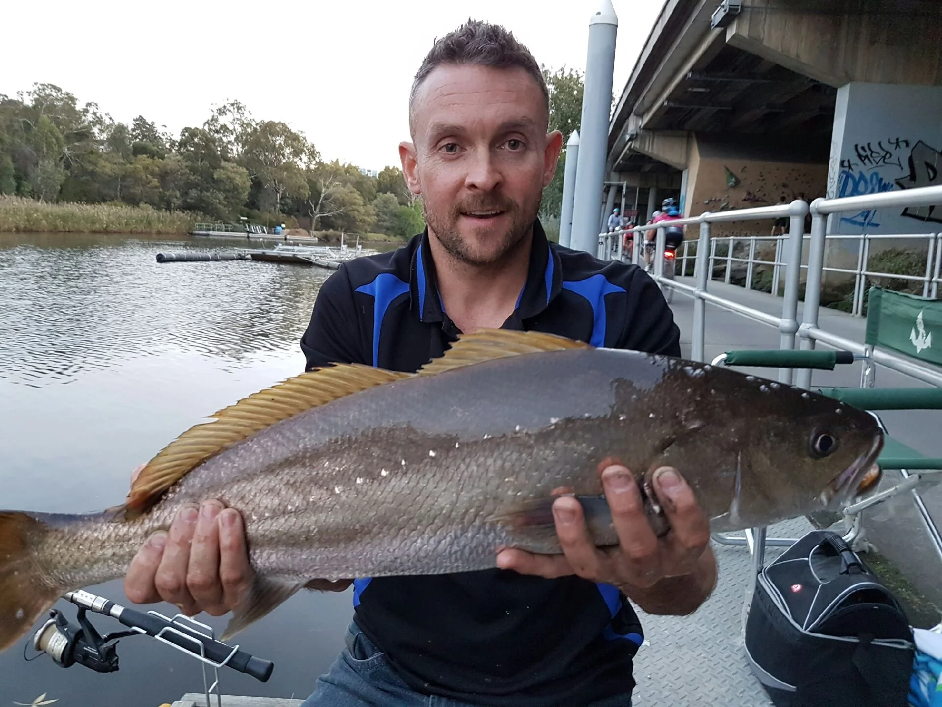 Man holding a large fish near a riverside, with a bridge and cyclists in the background.