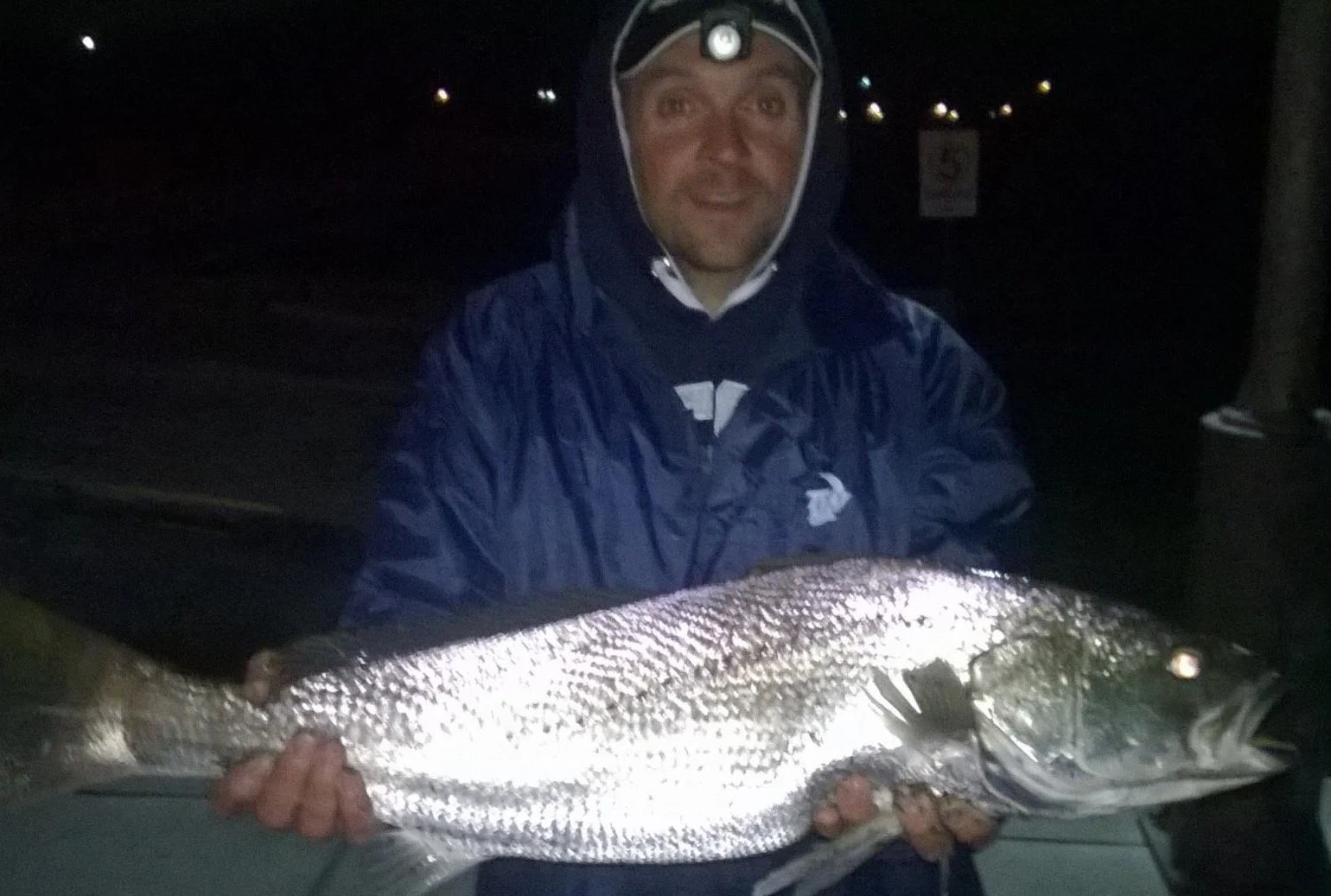 Person in dark clothing holding a large fish at night, with waterproof gear and a headlamp on their forehead.