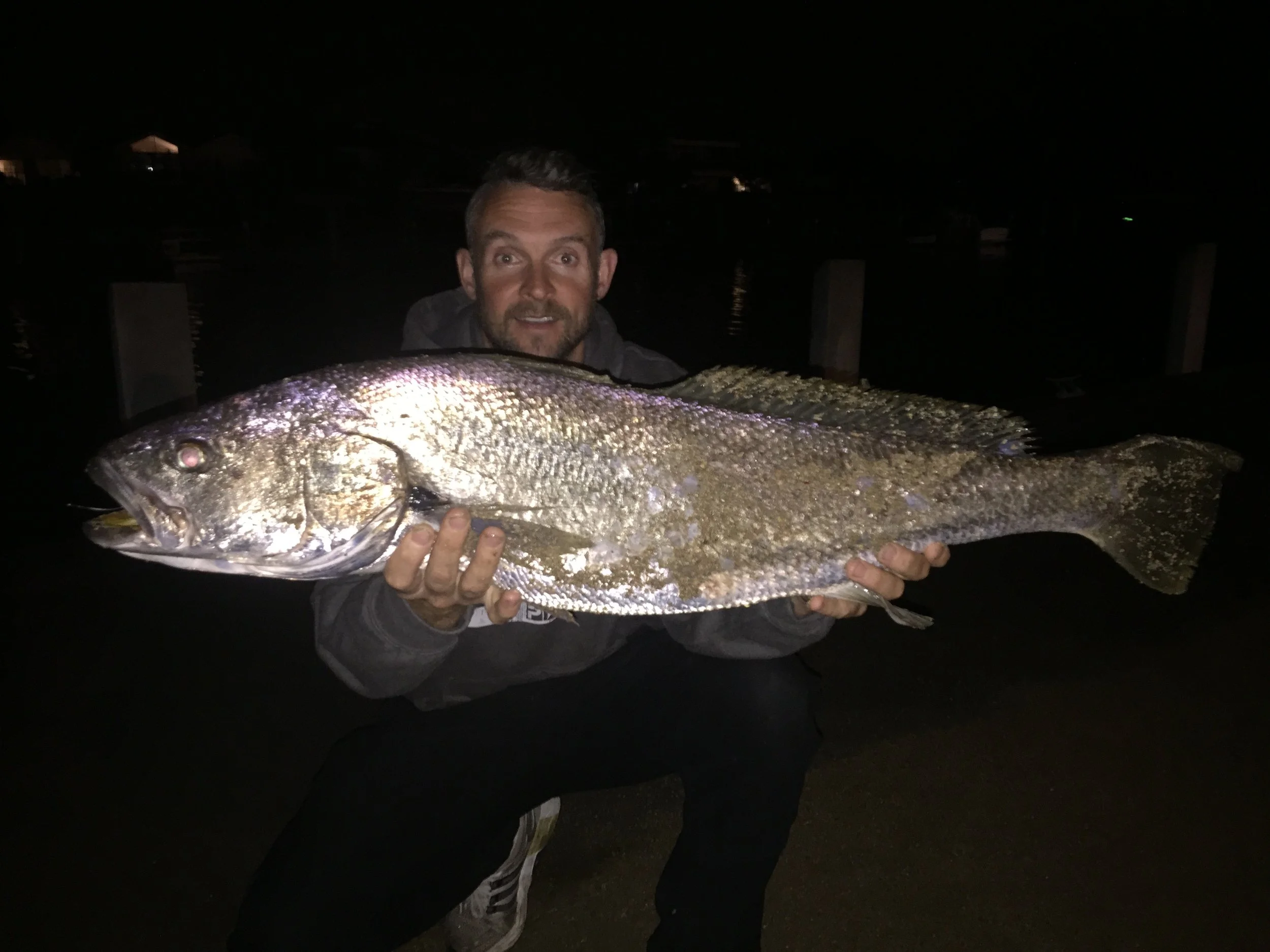 A man holding a large fish at night near the water, with a dark sky and faint lights in the distance.