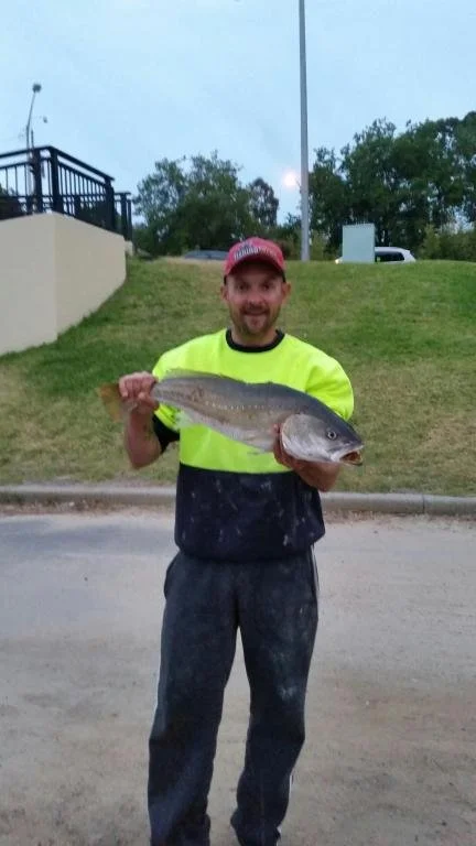 A smiling man wearing a red cap, neon yellow shirt, and dark pants is holding a large fish outdoors on a sidewalk, with grass, trees, and a blue sky in the background.