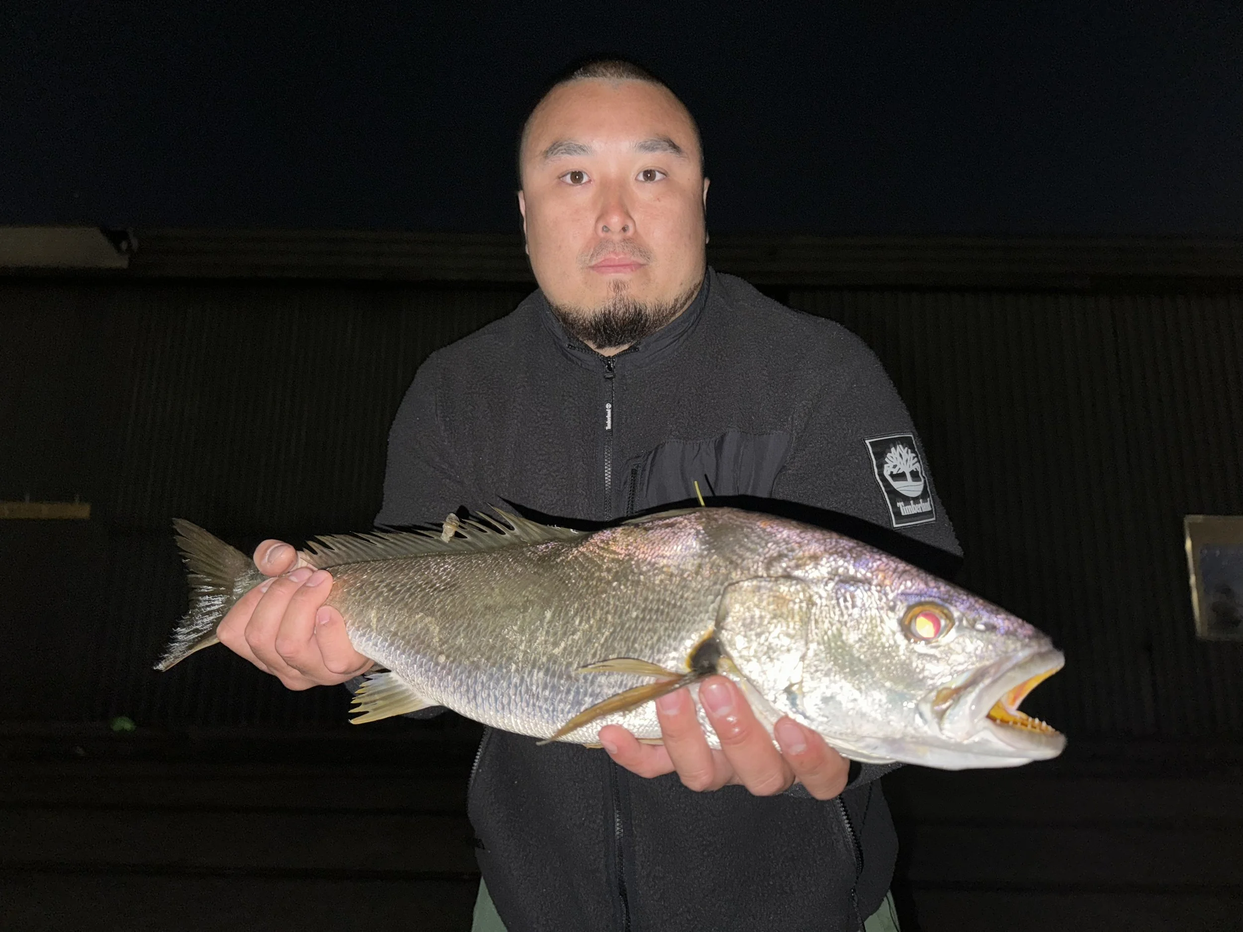 A person is holding a large fish, standing outdoors at night. The person is wearing a dark jacket and the background is dark.