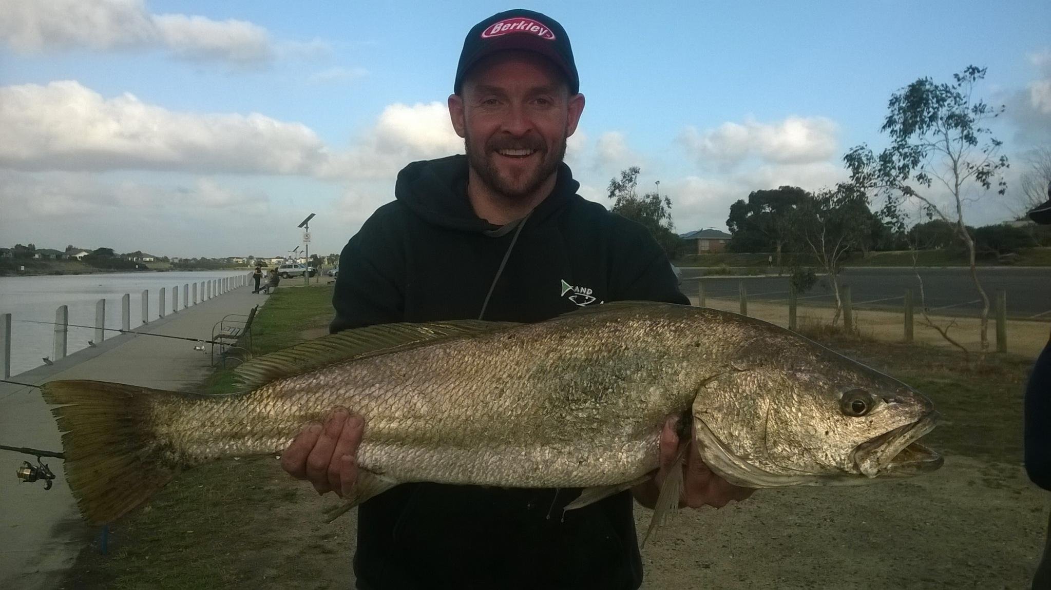 A man smiling while holding a large fish outdoors near a waterway, with trees and a cloudy sky in the background.