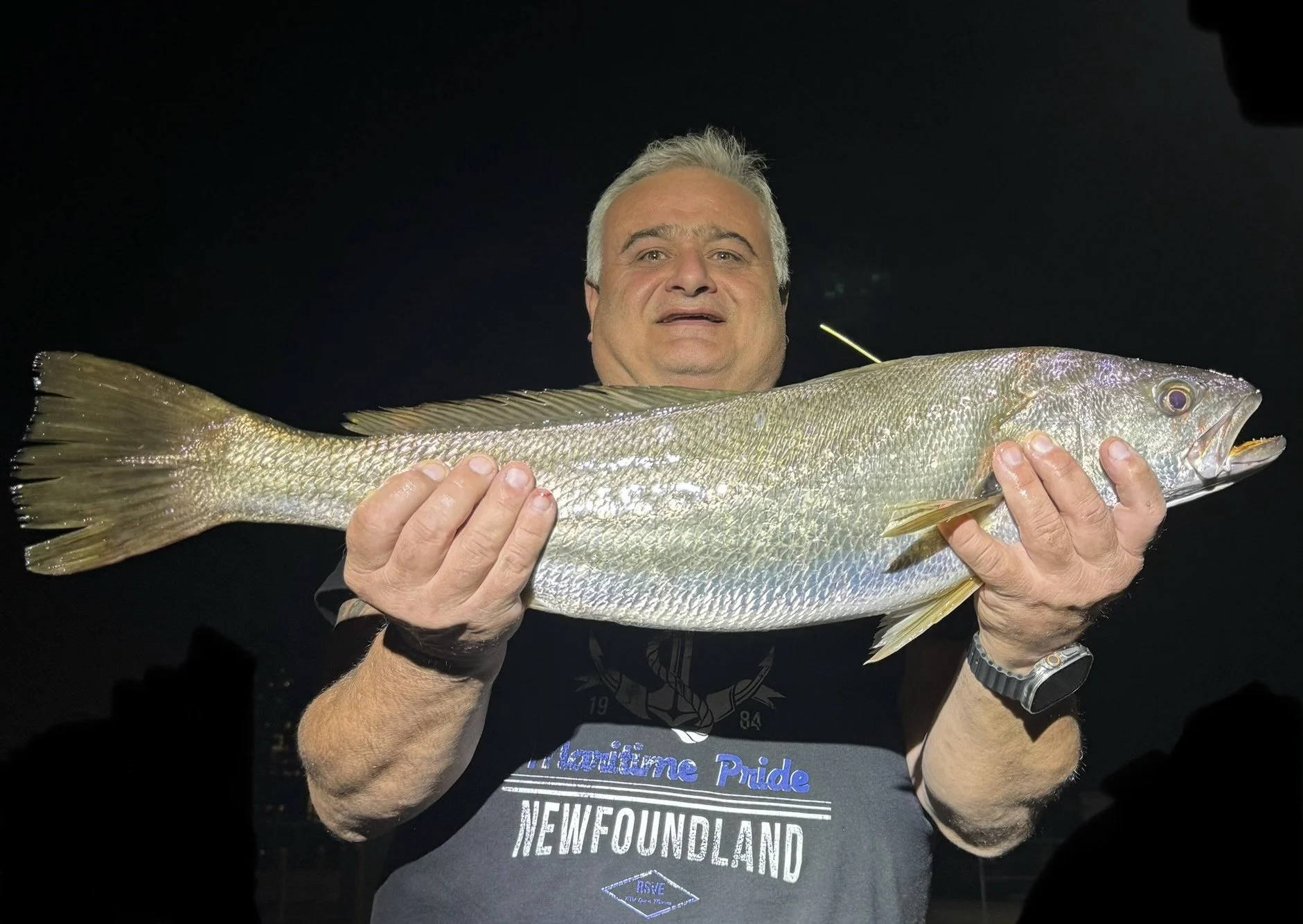 Man holding a large fish with both hands, dark background, wearing a grey t-shirt that says 'Newfoundland' with text indicating 'Labrador Pride'.
