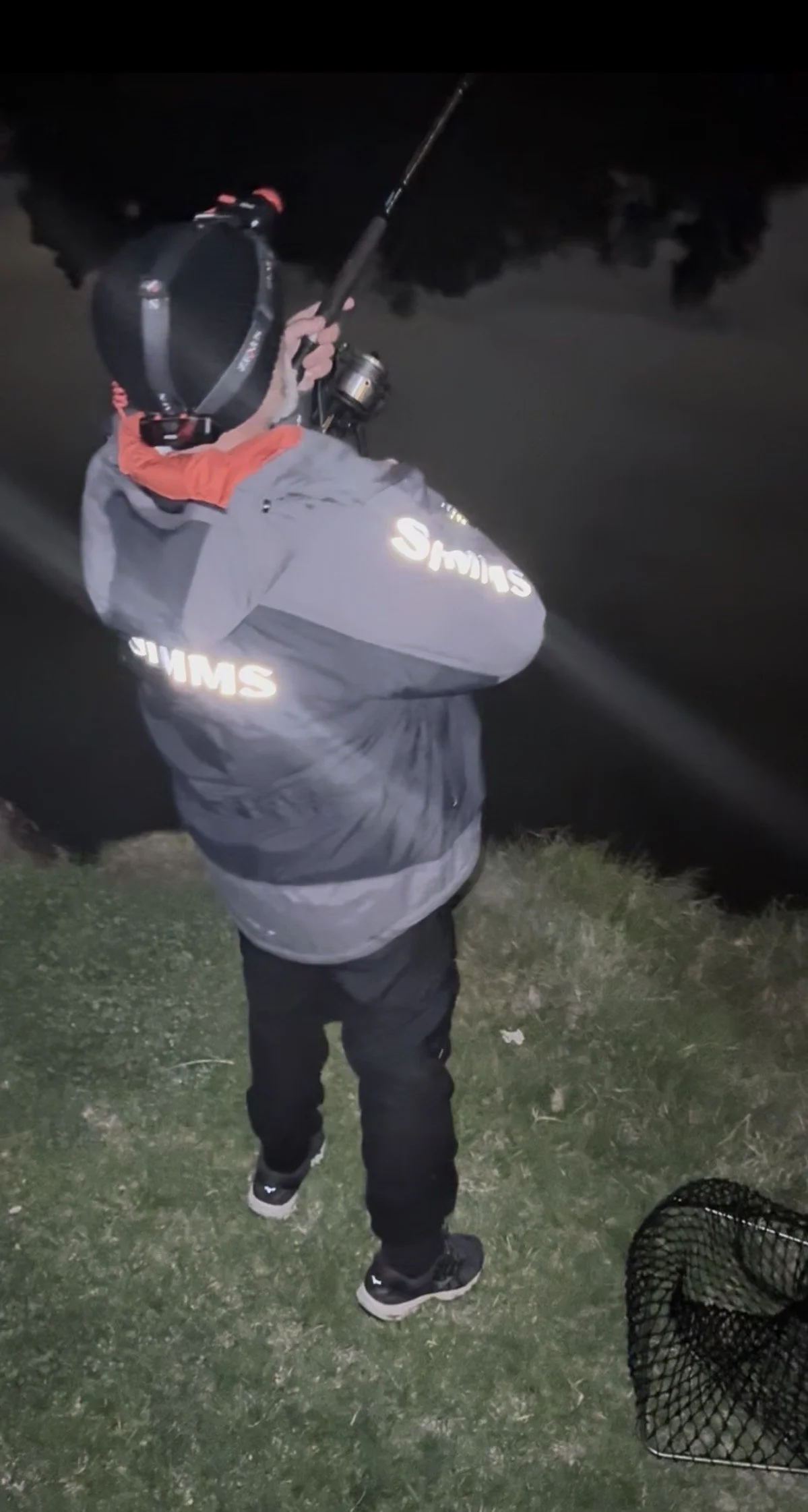 A person in dark clothing, wearing a jacket with reflective lettering, holding a fishing rod at night near a body of water, with a black net on the ground nearby.