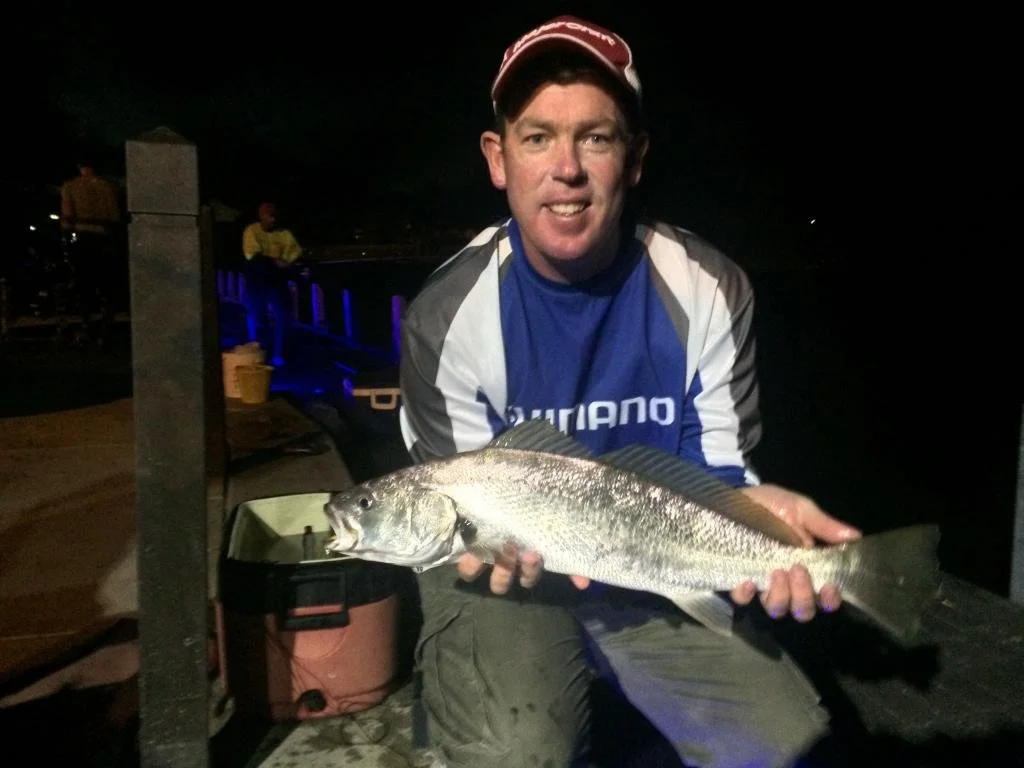 A man holding a large fish at night, wearing a blue and gray jacket and a red cap, with people and a dock in the background.