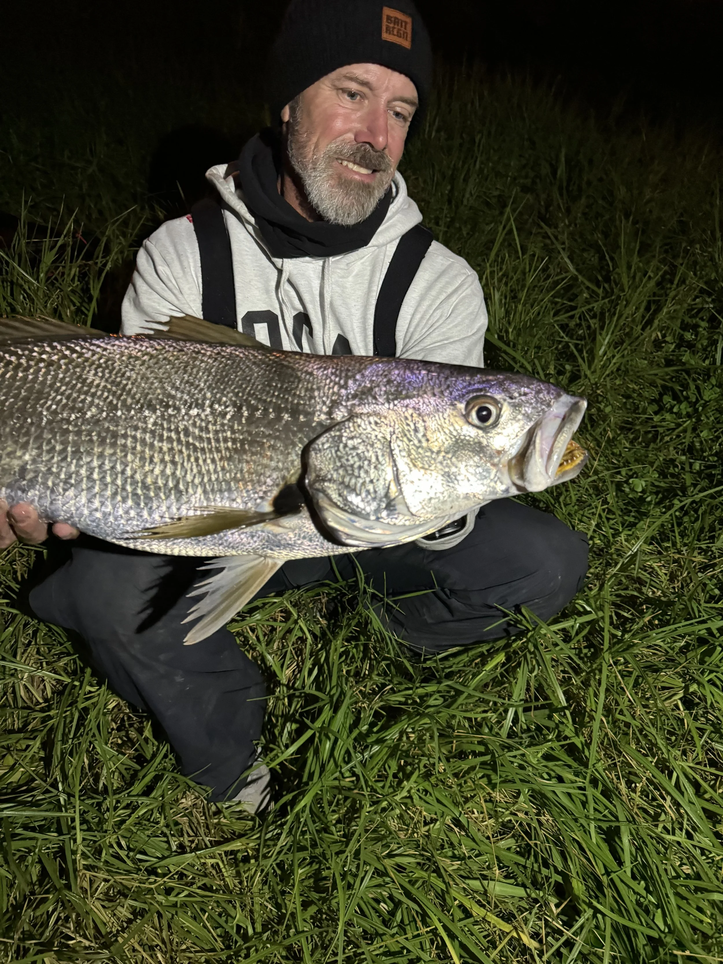 A man with a beard and a black beanie holding a large fish in tall green grass at night.