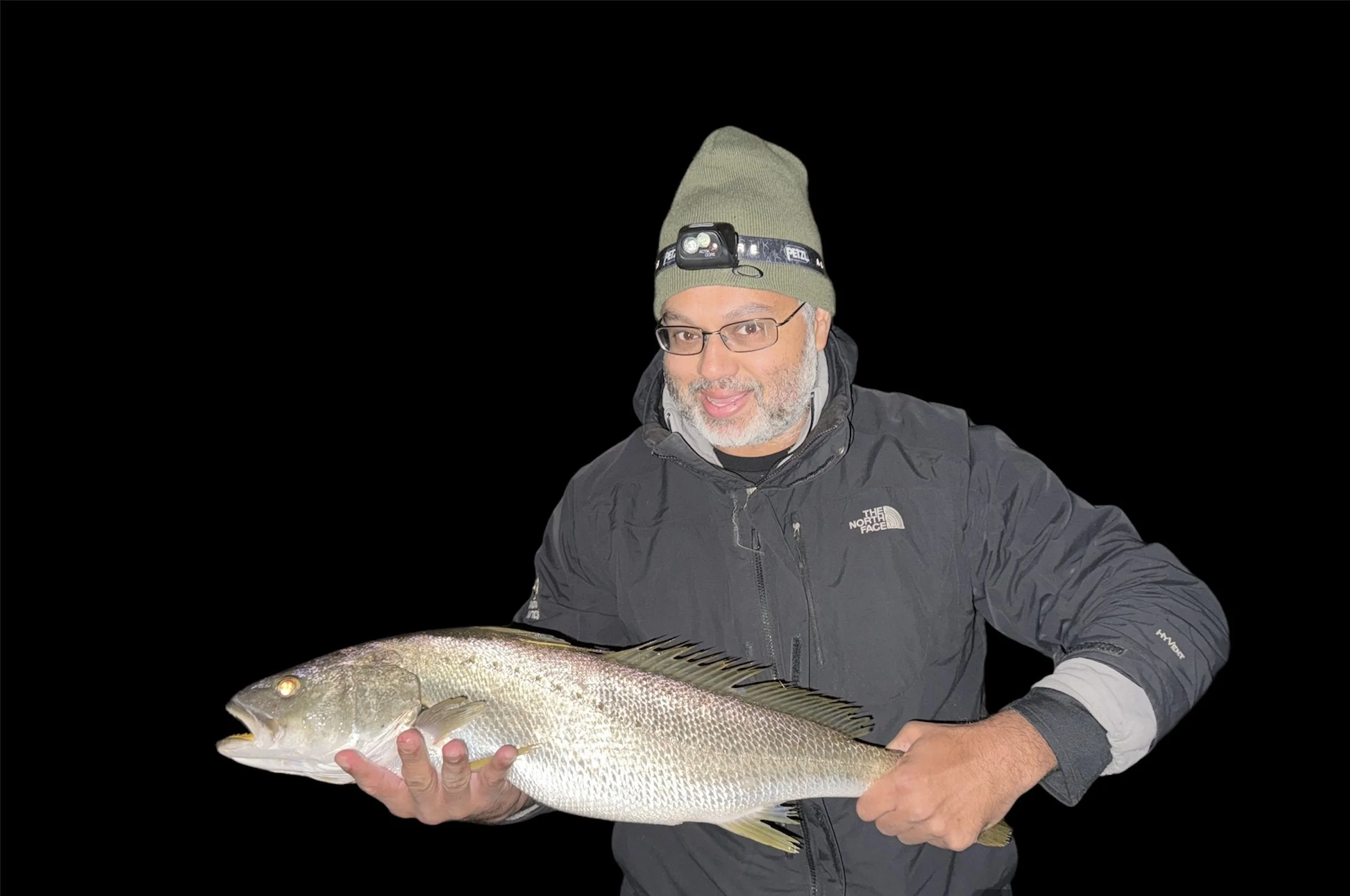 A man wearing glasses, a green beanie with a headlamp, and a black North Face jacket, holding a large fish against a black background.